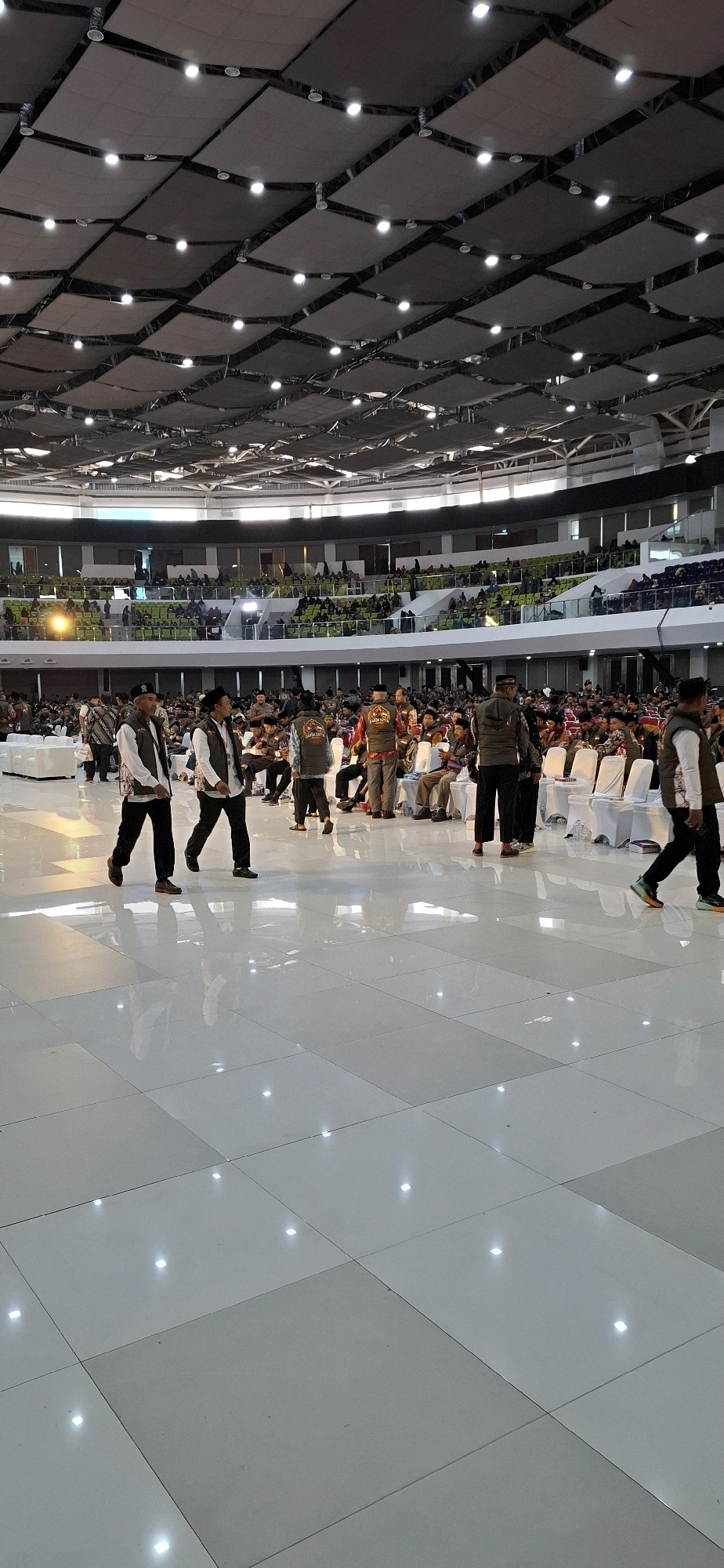 The image depicts an indoor event held at the Blulukan Sports Hall in Blulukan, Indonesia. The hall features a high ceiling with a dark, modular fabric structure interspersed with numerous spotlights. The floor is a polished, light-colored tile, reflecting the ambient light.
In the foreground, several men are walking across the polished floor. They are dressed in traditional attire, including vests over shirts and headwear, suggesting a formal or cultural gathering. Two men in the immediate foreground are walking towards the left, one with a black vest and white shirt, the other with a white vest and black shirt. Further back, other individuals are seen standing or walking, some wearing similar vests with various insignia.
The mid-ground reveals rows of white chairs arranged for an audience. Behind the chairs, spectators are seated in tiered seating that curves around the space. The seating areas are predominantly green. Further in the background, more spectators can be seen in the upper levels of the hall.
The overall atmosphere appears to be that of a large public event, possibly a conference, religious gathering, or cultural festival, taking place within a modern sports arena. The lighting is bright, illuminating the expansive space and highlighting the reflective floor.