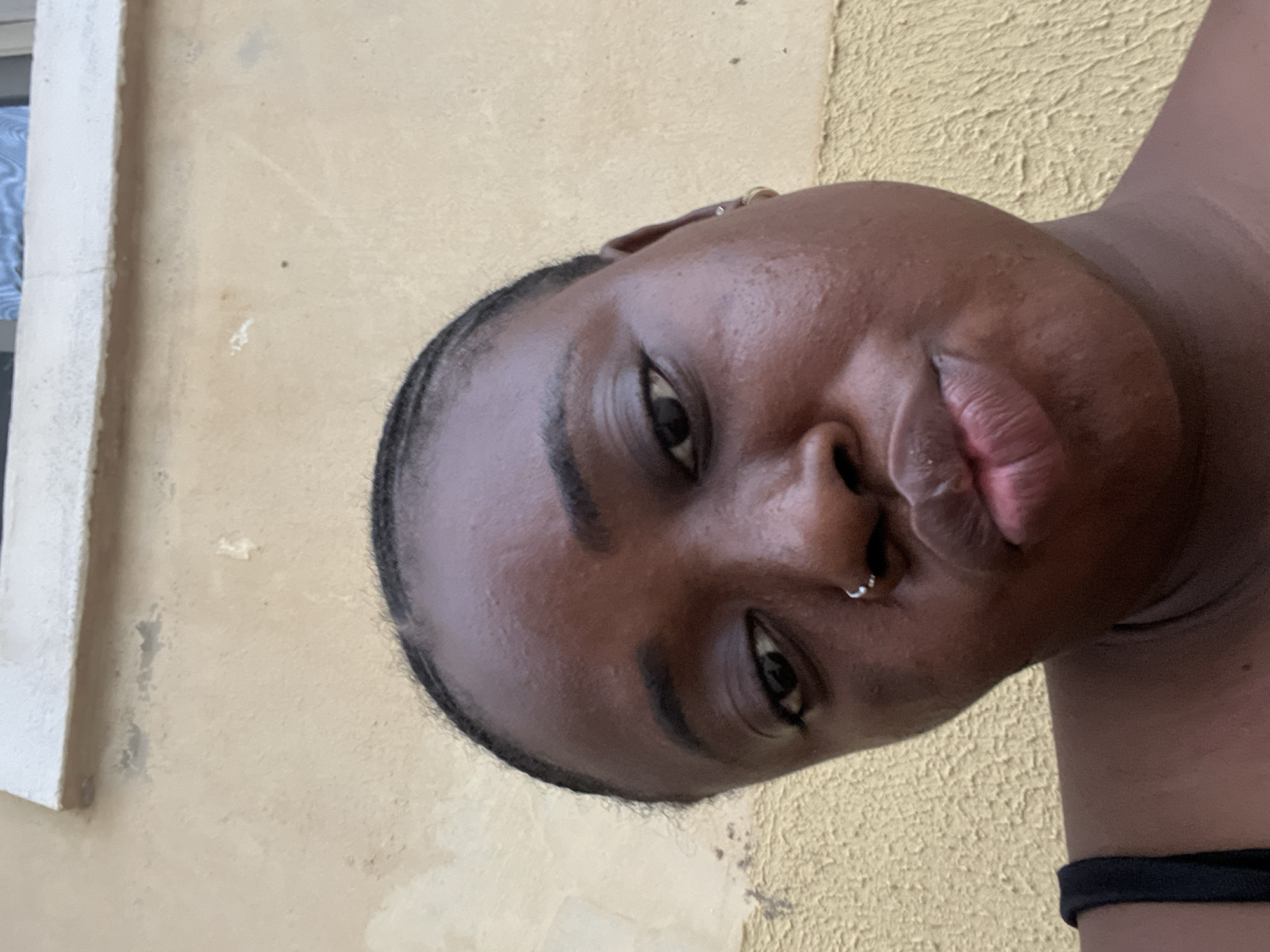 A medium shot of a Black woman with dark skin, a nose ring, and earrings, against a yellow textured wall and a white window frame. She is wearing a black choker around her neck. The location is likely in Abuja, Nigeria.