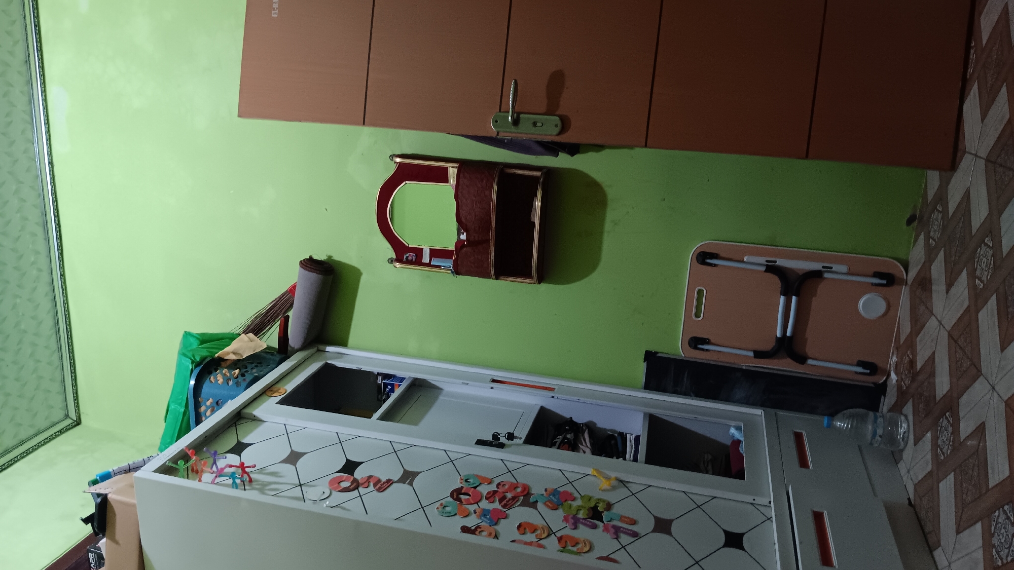 The image shows a corner of a room. A white wardrobe with decorative stickers is on the left. Inside the wardrobe, clothes and other items are visible. On top of the wardrobe, there are some items including a blue plastic basket, green plastic sheeting, a broom, and a rolled-up mat. To the right of the wardrobe, there is a small wall-mounted shelf with a mirror and compartments, decorated with red and gold trim. Further to the right is a brown wooden door with a silver handle. In front of the wardrobe and door, a folding laptop table with a light brown surface and white legs is placed on the floor. A plastic bottle of water is on the floor next to the wardrobe. The walls are painted light green, and the ceiling is also green with a patterned texture and a decorative border. The floor is tiled with a geometric pattern in brown and beige. The overall scene suggests a domestic interior in Sonobekel, Indonesia.