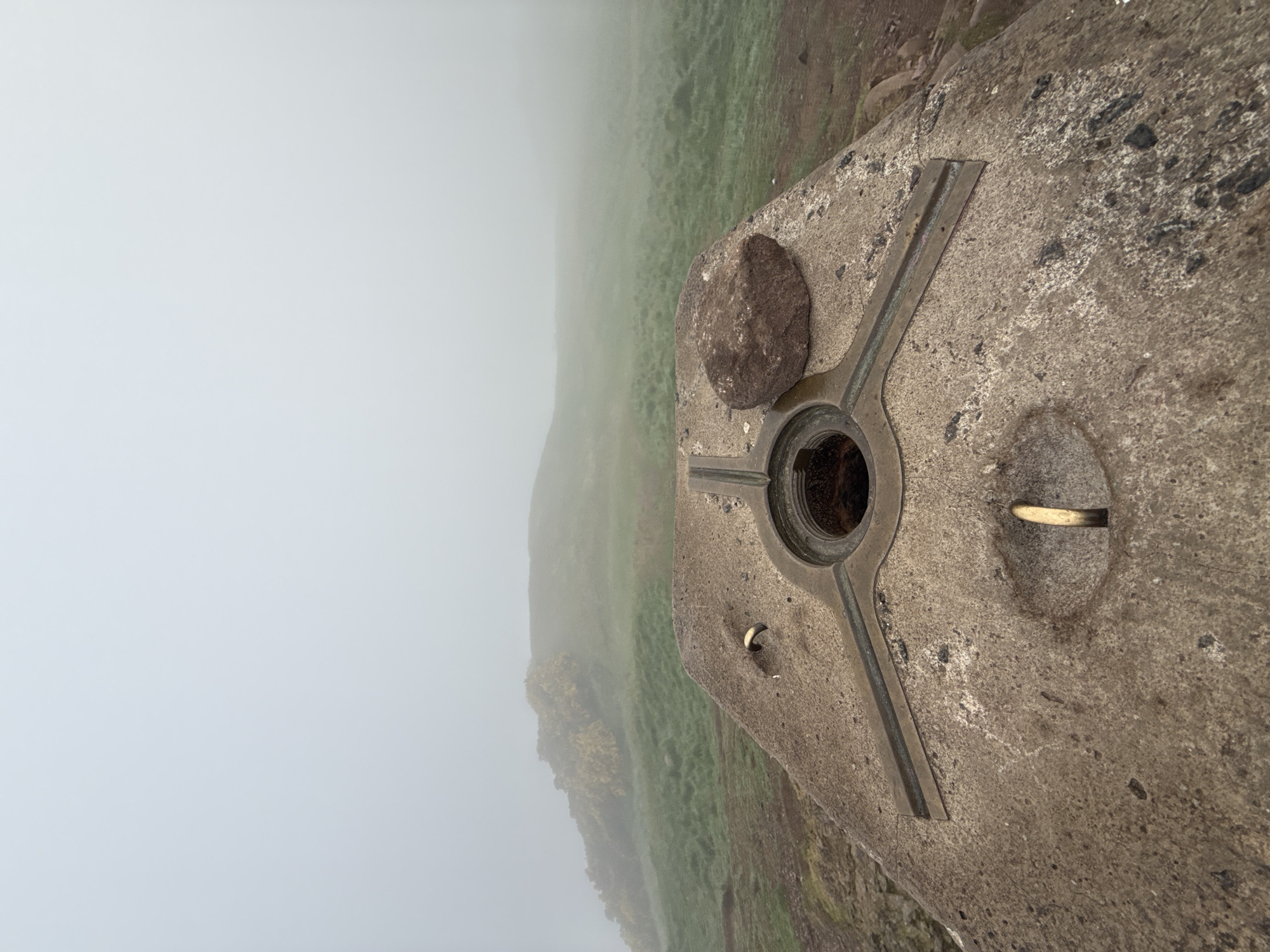 The image is oriented vertically (rotated 90 degrees clockwise), depicting a concrete survey marker, commonly known as a trig point, situated on an elevated outdoor landscape. The scene is dominated by dense fog or clouds that obscure the distant view.
The primary subject is a weathered, light grey concrete structure. It features a central circular metal fitting, dark in color, with a recessed center, designed for mounting surveying instruments. Three thin, dark metal strips extend outwards from this central fitting, embedded into the concrete surface. To the right of the central fitting, within another circular depression in the concrete, a small, bent, brass-colored metal object, resembling a hook or pin, is inserted. To the left of the central fitting, an irregular-shaped, dark brown rock rests on the concrete surface.
The background shows a rolling landscape covered in green and brown vegetation, likely grass and some scrub, fading into a thick, white layer of fog that occupies the majority of the left and upper parts of the image. The pervasive fog creates a sense of isolation and mystery, limiting visibility and diffusing the natural light. No people are visible, and there is no discernible text on the concrete marker or in the environment. The weather is clearly foggy or heavily overcast, making it difficult to ascertain the exact time of day, though it appears to be daylight. The location cues point to a mountain or hill summit due to the presence of the trig point and the expansive, although obscured, vista.
