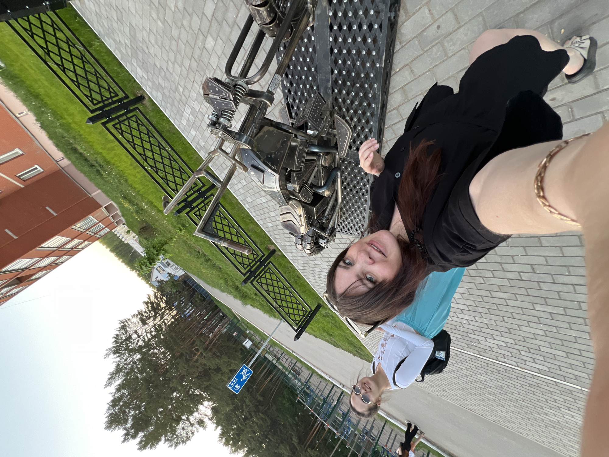 A photograph taken in Chaikovsky, Russia, shows a woman posing with a metal sculpture of a motorcycle. The woman is wearing a black dress and stands next to the sculpture. To the left of the sculpture is a fenced green space with a red brick building in the background. A second woman stands further away, wearing sunglasses and a white top. A sign with a wheelchair symbol is visible in the background. The scene is captured on a paved surface.