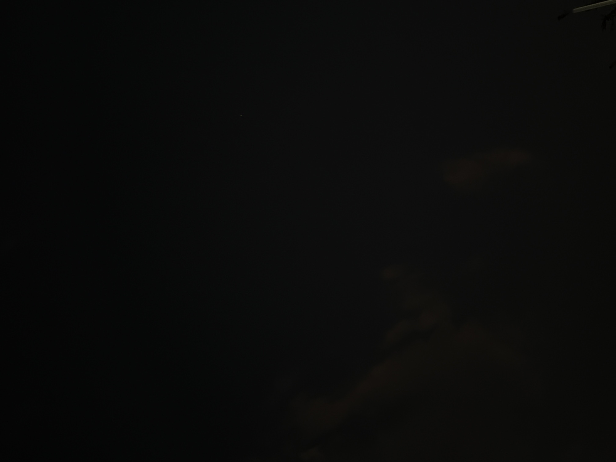 The image depicts a night scene, possibly in Cuautla, Mexico, given the request. The majority of the frame is dark, suggesting low light conditions. There is a single, small light source visible near the center of the upper portion of the frame. In the lower-right quadrant, shapes suggest shadows that might be clouds. At the top-right edge are shapes that suggest a pole with tree branches. Overall, the image is mostly dark.