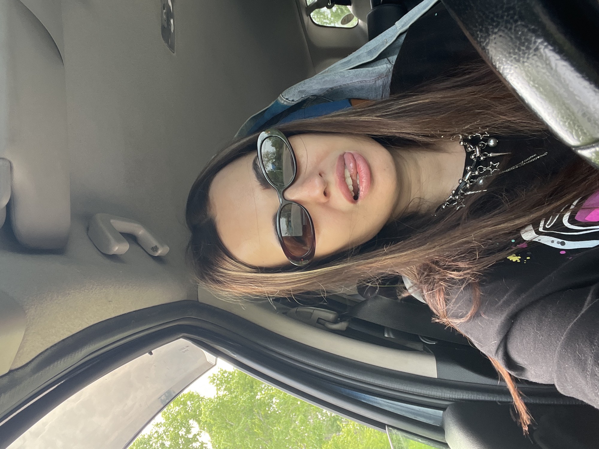 A young adult woman is in the driver's seat of a car. She is wearing sunglasses, a black shirt, and a metal chain necklace. Her hair is long and brown. She is sticking her tongue out. The car is parked, and trees are visible through the window. The location is Unknown, Unknown.