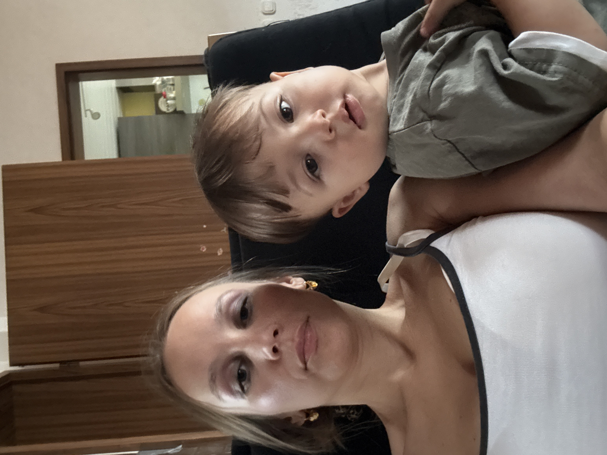 The image is a selfie of a woman and a young child taken indoors. The woman, on the left, has light brown hair and is wearing a white tank top. She has makeup on, including eyeshadow and lipstick. She is wearing gold hoop earrings. The child, on the right, has brown hair and is wearing a gray t-shirt. They are both looking towards the camera. The background appears to be a living room, with wooden furniture and a doorway visible in the reflection of a mirror. There is no information in the image to suggest this location is in Überherrn, Germany.