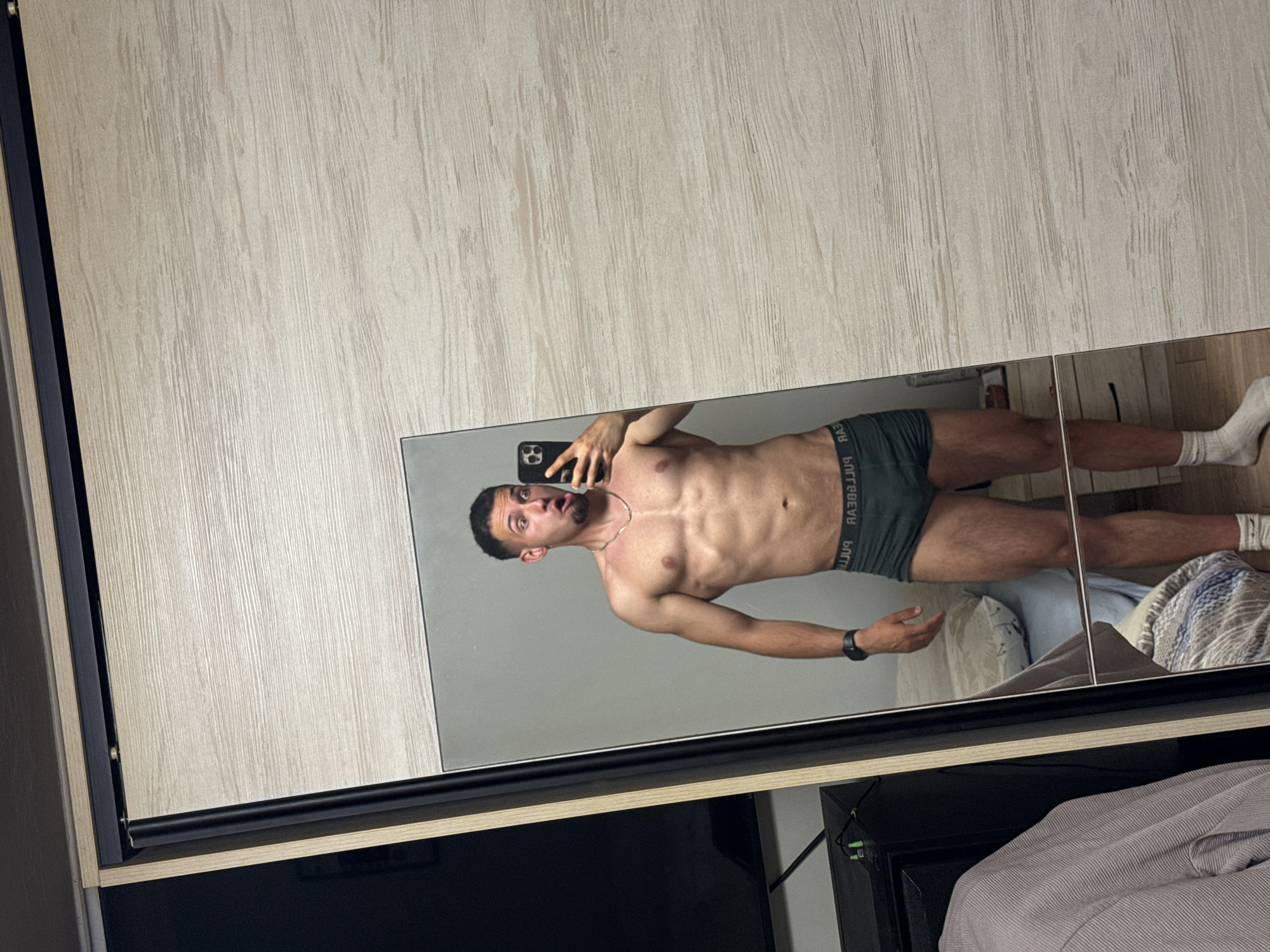 A man in underwear takes a mirror selfie in what appears to be a bedroom. The background shows a bed and a wardrobe. The exact location in Shumen, Bulgaria is undeterminable from this image.