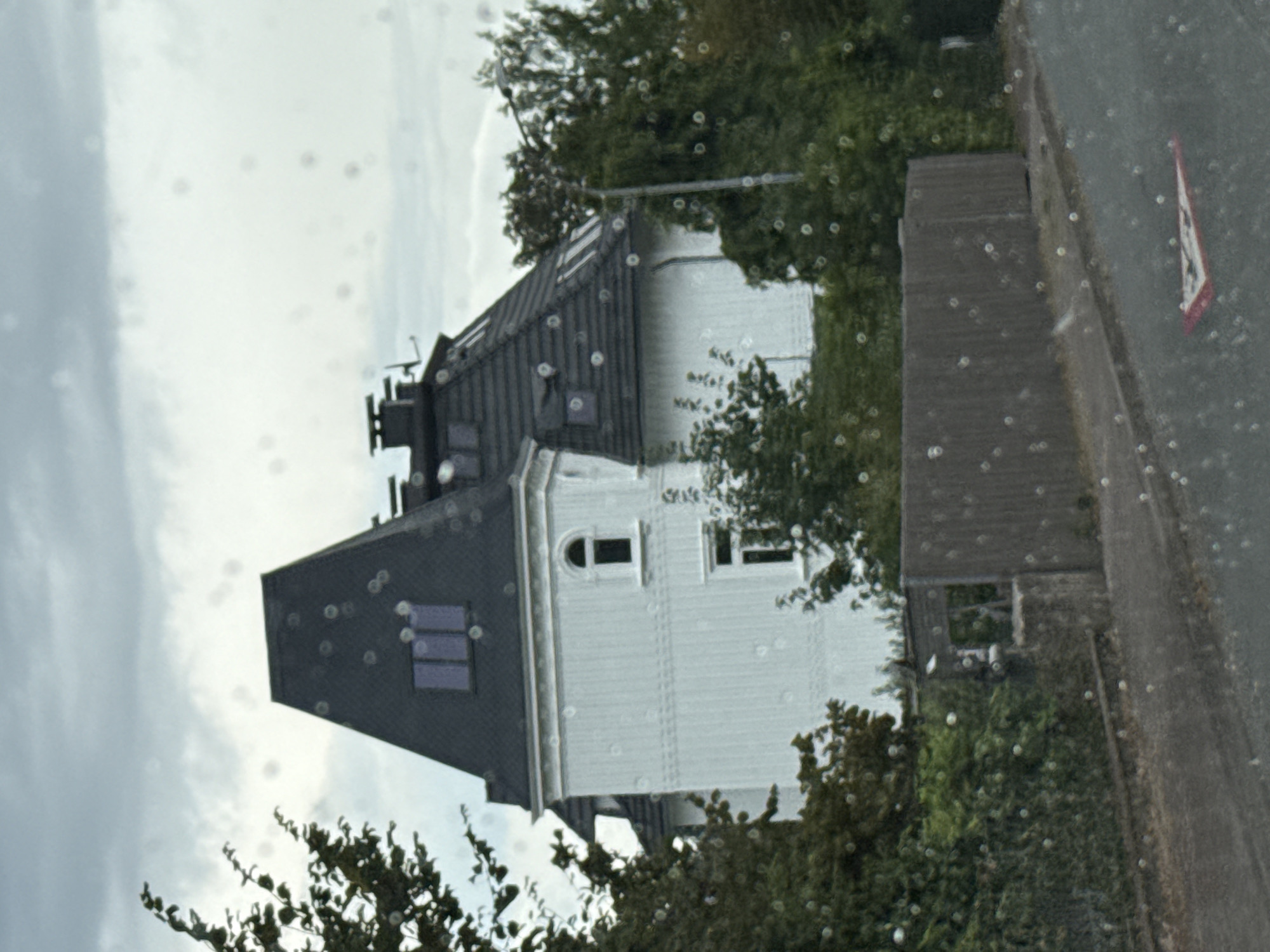 This is a medium-shot, eye-level, outdoor, full-color photograph taken from a vehicle. The image shows a light-colored, two-story house with a dark, mansard-style roof and several dormer windows. The house is situated behind a wooden fence and surrounded by lush green trees and bushes. A paved road with a warning sign is visible in the foreground. The sky is overcast with clouds. The scene appears to be in a residential area of Bad Sachsa, Germany.