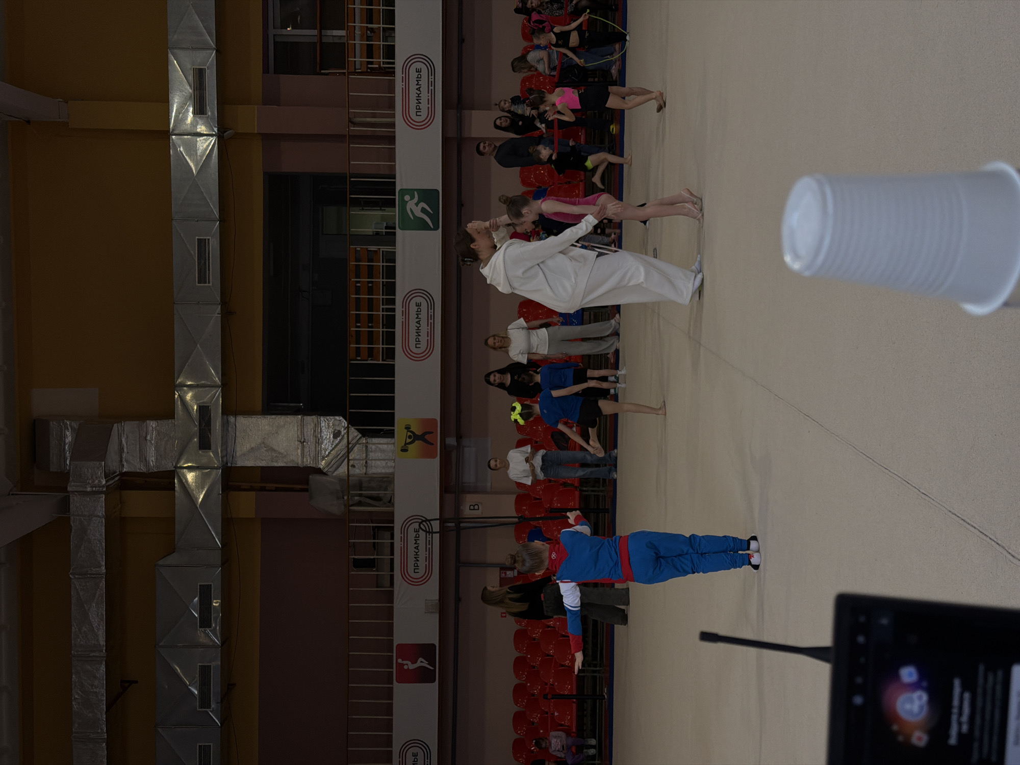 The image shows the interior of a gymnasium, likely in Perm, Russia, judging by the "ПРИКАМЬЕ" signage. There are groups of people standing and interacting, including individuals wearing athletic attire. The environment includes rows of red chairs lining the walls and visible ventilation systems on the ceiling. Other elements such as a plastic cup and an electronic device are positioned in the foreground.