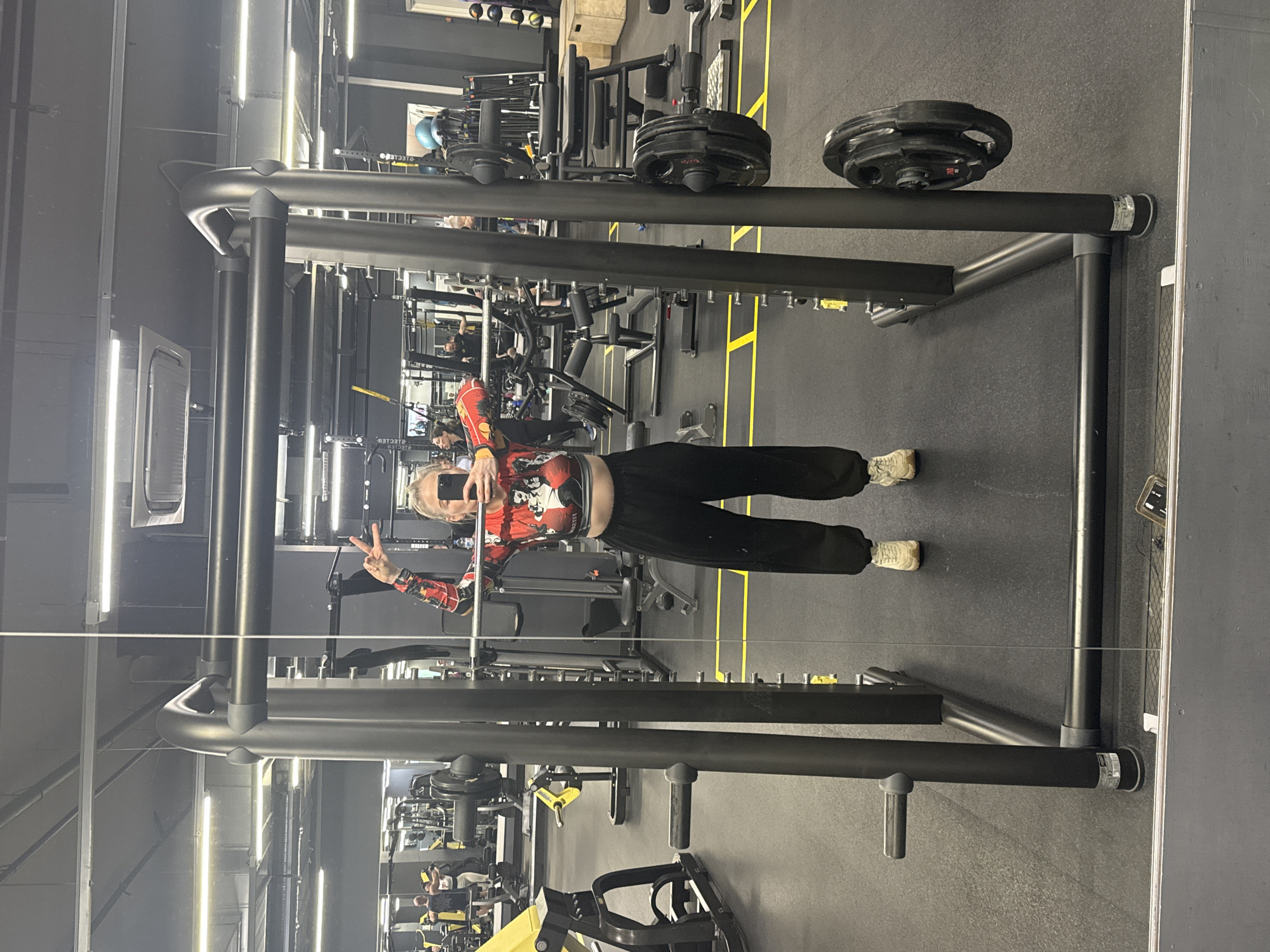 A person is taking a selfie in a gym. The gym has various workout machines and equipment. The person is wearing black pants, a red and black shirt, and white sneakers. They are holding a phone in their left hand and making a peace sign with their right hand. The gym has a black floor with yellow lines. There are black weights stacked on a squat rack. The lighting in the gym is bright. The location is not in Saint Petersburg, Russia.