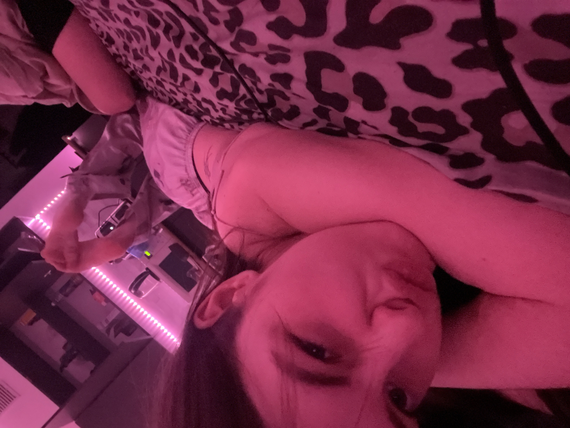 A person is lying in bed, with the face close to the camera. The bedding has a black and white animal print. A bedside table with a row of LED strip lights is visible, featuring items such as a computer screen, a container with what appears to be charging cables, and other small items. The photo is dominated by pinkish-red lighting. The location in the city of Saint Petersburg, Russia is not possible to determine from the image.