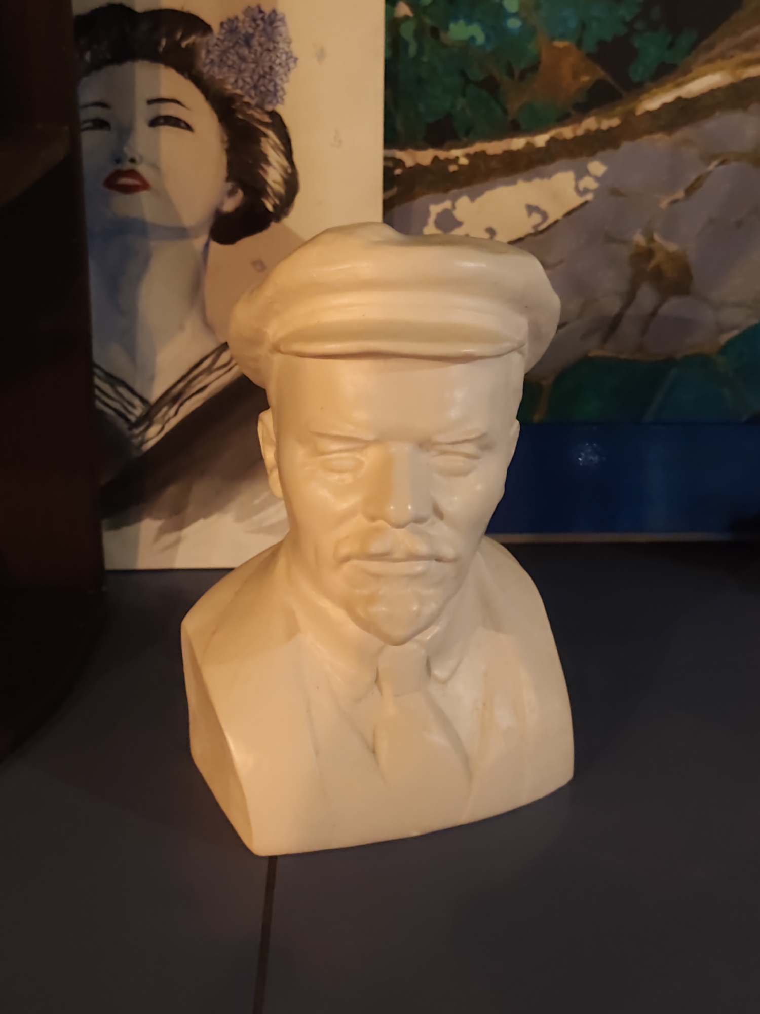 The image shows a bust of Vladimir Lenin, made of a light-colored material, likely plaster or resin. Lenin is depicted wearing a cap and a shirt with a tie. The bust is positioned in the foreground. Behind it, to the left, is a painting or print of a woman with traditional Japanese hairstyle and makeup. To the right of the bust, a portion of another artwork with abstract shapes in green, brown, and blue colors is visible. The background appears to be a wall or display, with a dark blue element at the bottom right. The overall setting suggests an indoor display of art or collectibles. The image does not provide any information about the city of Sochi, Russia.