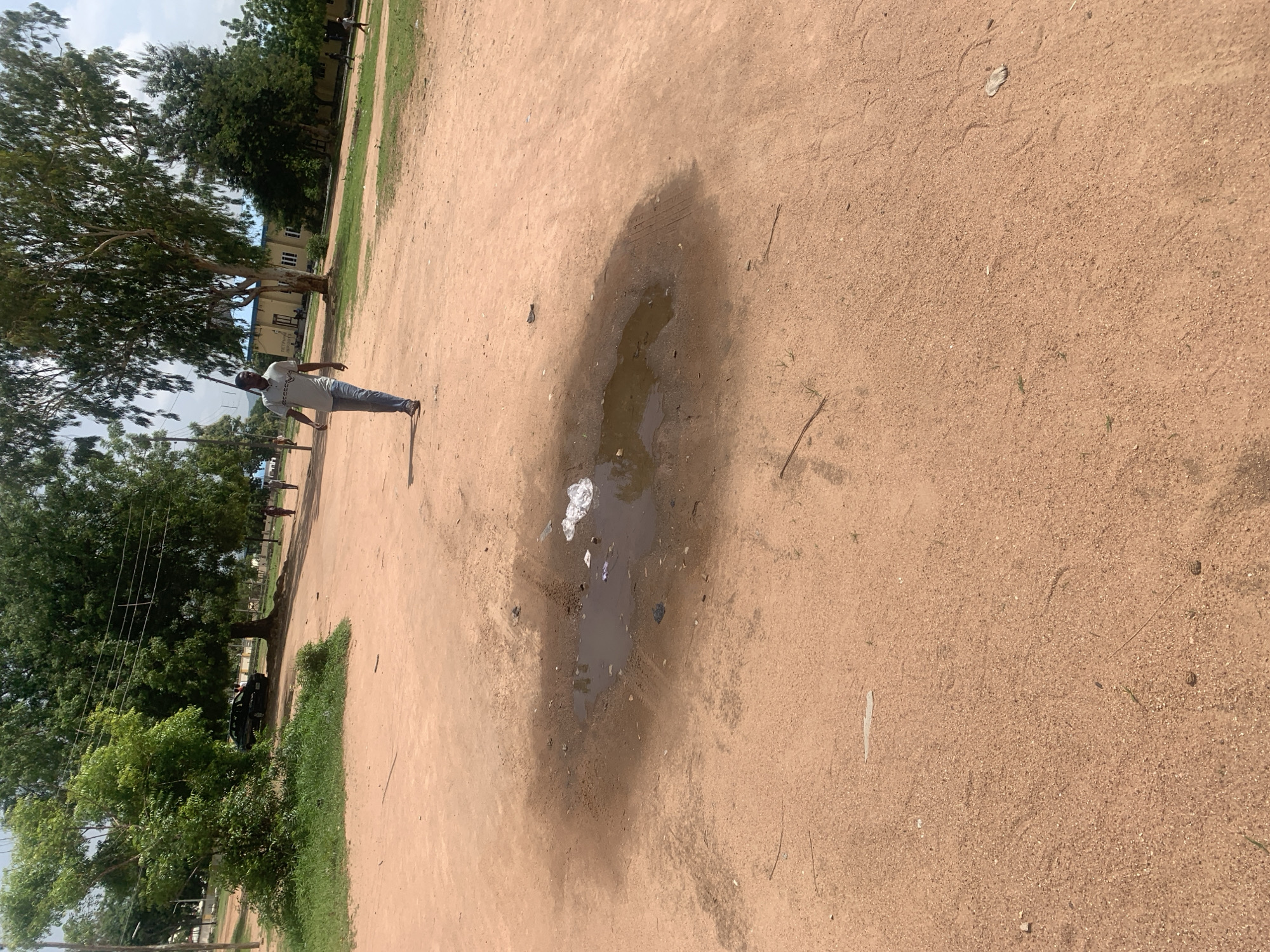 In Jalingo, Nigeria, a man walks across a reddish-brown dirt road. The road is uneven and shows signs of recent rain, with a large puddle of muddy water in the foreground. Scattered around the puddle are pieces of white plastic trash. In the background, a line of trees with green foliage borders the road, and behind them, a light-colored building with a blue roof stands under a cloudy sky. The scene appears to be a typical outdoor area in a Nigerian city, possibly a campus or residential neighborhood.
