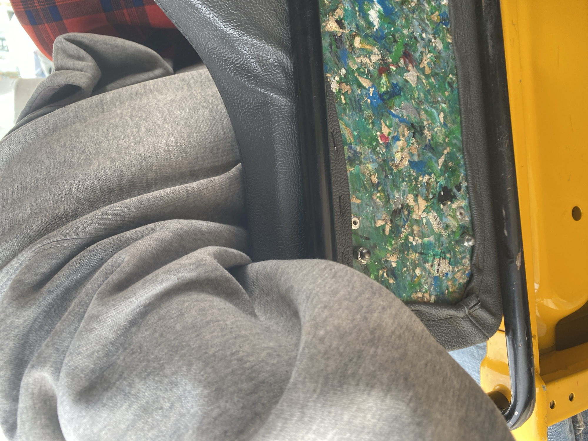 The image shows a close-up view of a person sitting inside what appears to be a yellow vehicle, possibly a tuk-tuk or a similar mode of transport, given the context of Jalingo, Nigeria. The person is wearing a grey hooded sweatshirt, and the fabric of the sweatshirt is prominently featured, with its folds and texture visible. A glimpse of a red and blue plaid shirt is visible beneath the sweatshirt.
The interior of the vehicle reveals a section with a black, textured surface, likely a seat or a panel. Adjacent to this is a decorative panel made of colorful, recycled materials, including pieces of blue, green, white, and red, embedded in a lighter matrix. This panel is secured with visible bolts. A black metal bar is also present, likely part of the vehicle's structure. The exterior of the vehicle is a bright yellow.
The image is an indoor shot, suggesting the vehicle is stationary or the lighting is controlled. There are no specific cues for time of day or weather. The focus is on the tactile textures of the clothing and the vehicle's interior. The scene is ordinary, depicting a moment of being seated within the vehicle. No text is visible in the image.