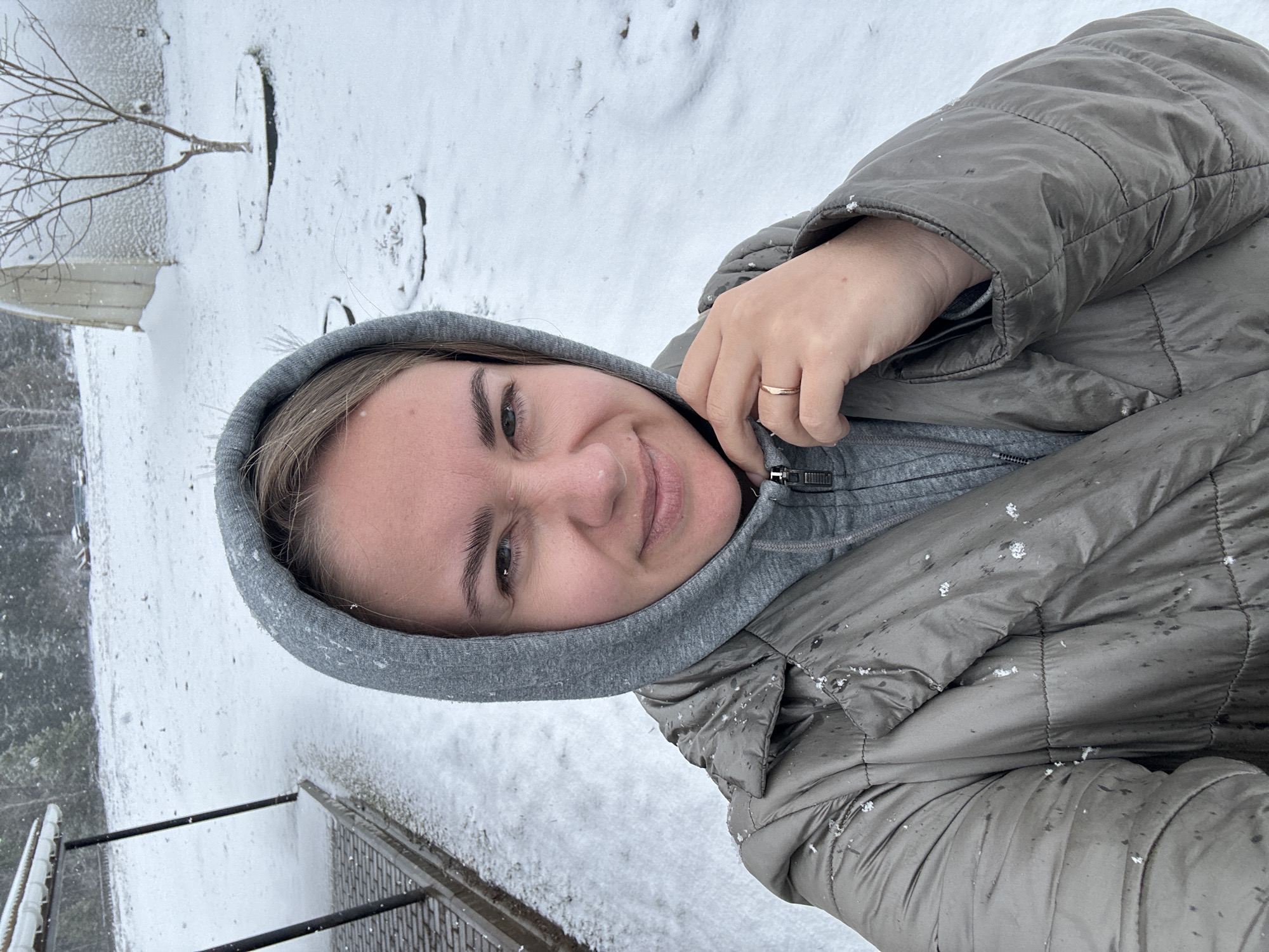 A young woman in a grey hooded jacket smiles into the camera. She is standing outside, in a snowy landscape. Snowflakes are visible on her jacket and in the background. A railing and what appears to be part of a building or structure are partially visible in the snow-covered background. The location is Уральское, Russia.