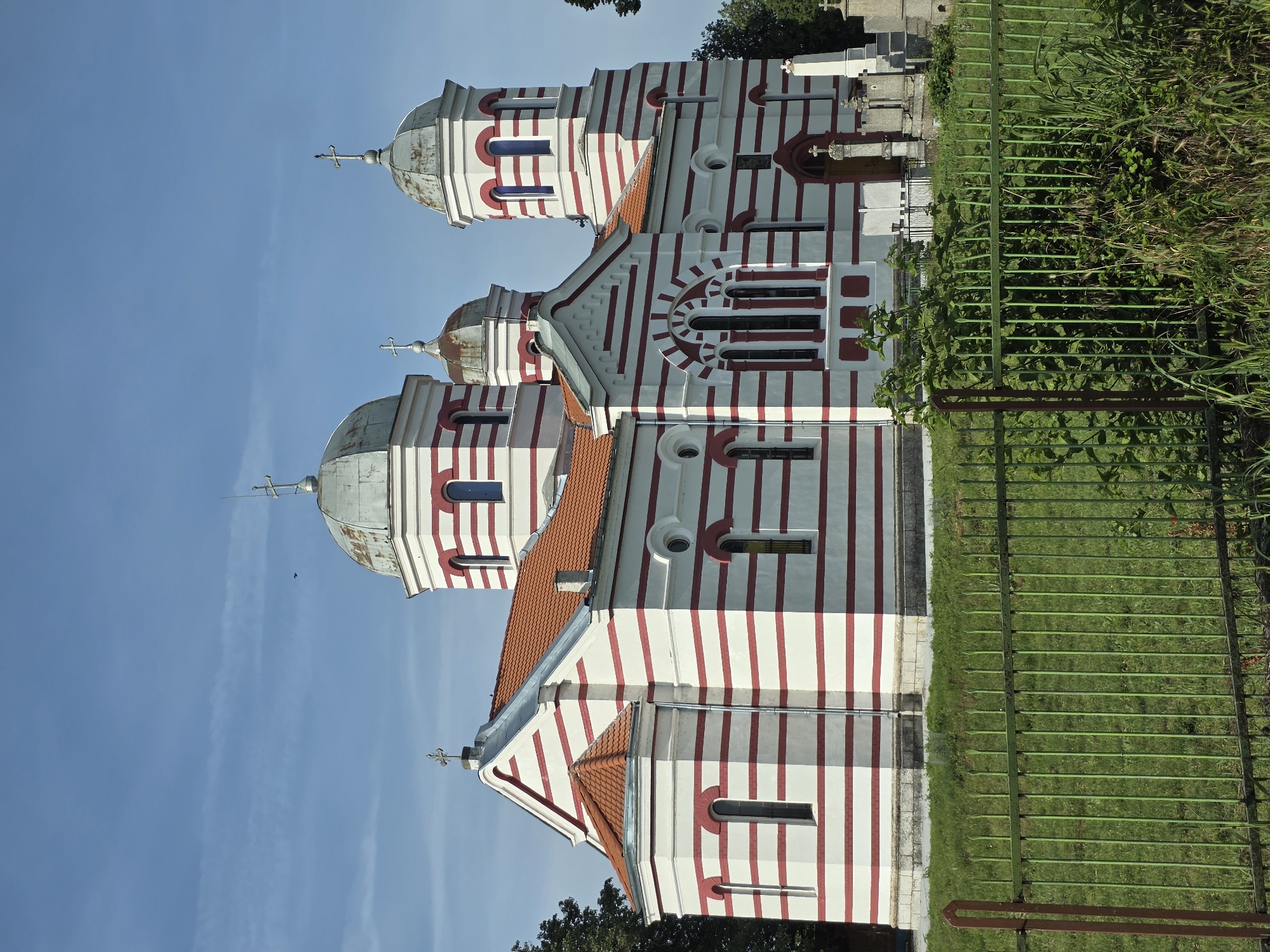 The image displays an exterior view of a distinctive church, characterized by its white facade with bold, vertical red stripes. The church features at least three visible domes, two larger ones in the foreground and one smaller dome further back, all topped with crosses. The domes exhibit a weathered, metallic grey appearance. The roof sections are covered with orange-red terracotta tiles. The building incorporates various window shapes, including arched, rectangular, and circular designs.
In the foreground, to the right of the church, a green metal fence runs vertically through the frame, separating the church property from an area of tall green grass and abundant foliage. Behind the fence, near the church's rear section, what appears to be a white tombstone or monument is partially visible, suggesting an adjacent cemetery. The sky above is a clear, bright blue, indicating a sunny day. The lighting is bright and even, typical of daytime conditions. No people or specific activities are depicted, and no visible text is present in the scene. The architectural style hints at an Eastern Orthodox church, possibly located in a rural or semi-rural setting.