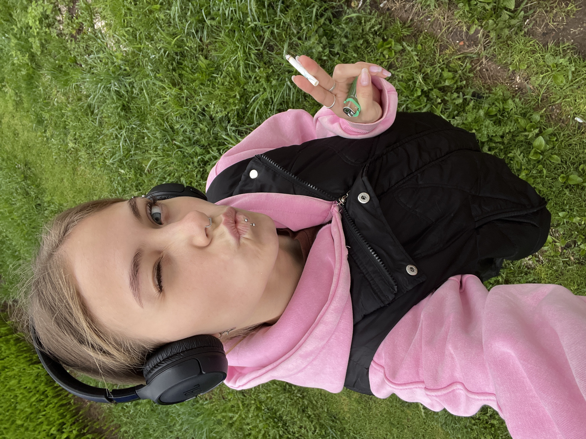 A young woman with light skin and blonde hair is lying on green grass. She is wearing a pink hoodie, a black vest, and black headphones around her neck. She has a piercing in her nose and two piercings on her lip. She is holding a lit cigarette in her right hand, and her lips are pursed in a kissing face. The location is a grassy area, potentially a park or field, in an unidentified location, not necessarily Фоки, Russia.
