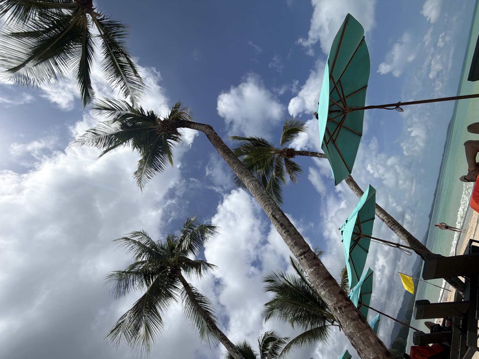 This is a low-angle, full-shot photograph of a tropical beach scene in Bang Muang, Thailand. The image is dominated by the bright blue sky, filled with scattered white clouds, and the fronds of several leaning palm trees that cast shadows on the sand. In the foreground, there are several turquoise beach umbrellas and lounge chairs, some occupied by unseen individuals. A yellow flag flies on a pole between the palm trees. Further back, a person stands in the shallow turquoise water near the shore, which is lined with mountains in the distance. The overall impression is one of a sunny, relaxed day at a seaside resort.