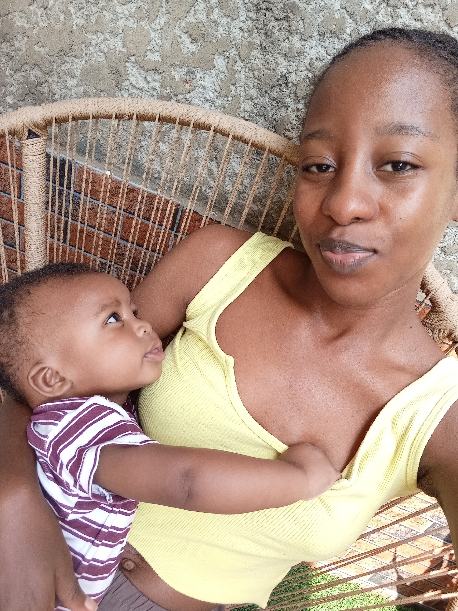 A young Black woman is holding a baby in her lap. She is wearing a yellow tank top. The baby is wearing a striped onesie. They are sitting in a rattan chair with a brick wall behind them. The woman is looking at the camera, and the baby is looking up at her. The lighting suggests it is daytime. The scene is outdoors.