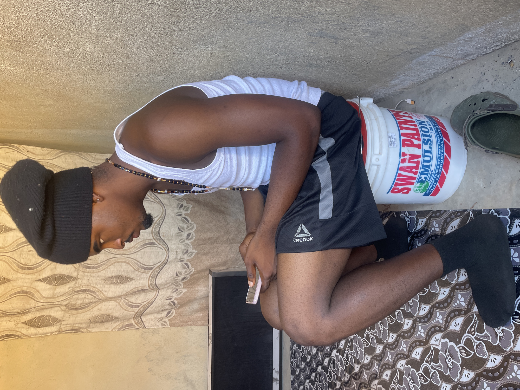 A man wearing a black beanie, white tank top, and black Reebok shorts is kneeling on a patterned mattress. He is holding a mobile phone in his hand. Next to him is a white bucket of SWAN PAINT emulsion and a green clog-style shoe. The scene is illuminated by sunlight. The location is possibly indoors, with patterned wall coverings visible. No specific details confirm the location as Durimi, Nigeria.