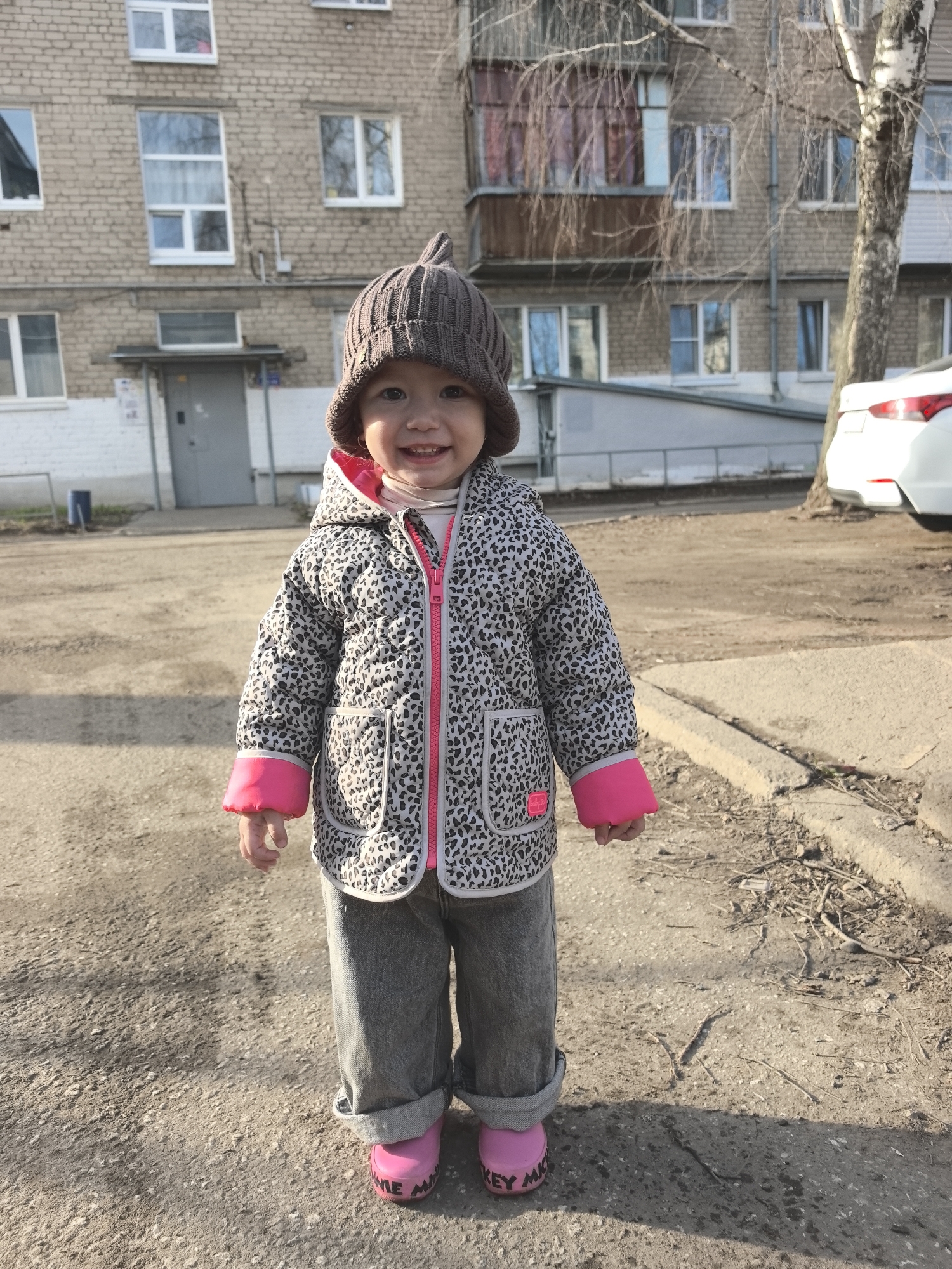 The image shows a young child standing on a cracked asphalt surface outdoors. The child is wearing a brown knitted hat, a gray and white patterned jacket with pink accents, gray jeans, and pink rubber boots. The child is smiling and looking directly at the camera. Behind the child is a multi-story brick building with multiple windows and balconies, and a birch tree. The back of a white car is partially visible to the right of the tree. The location is likely in a residential area of Chaikovsky, Russia, based on the style of the building and the surrounding environment.