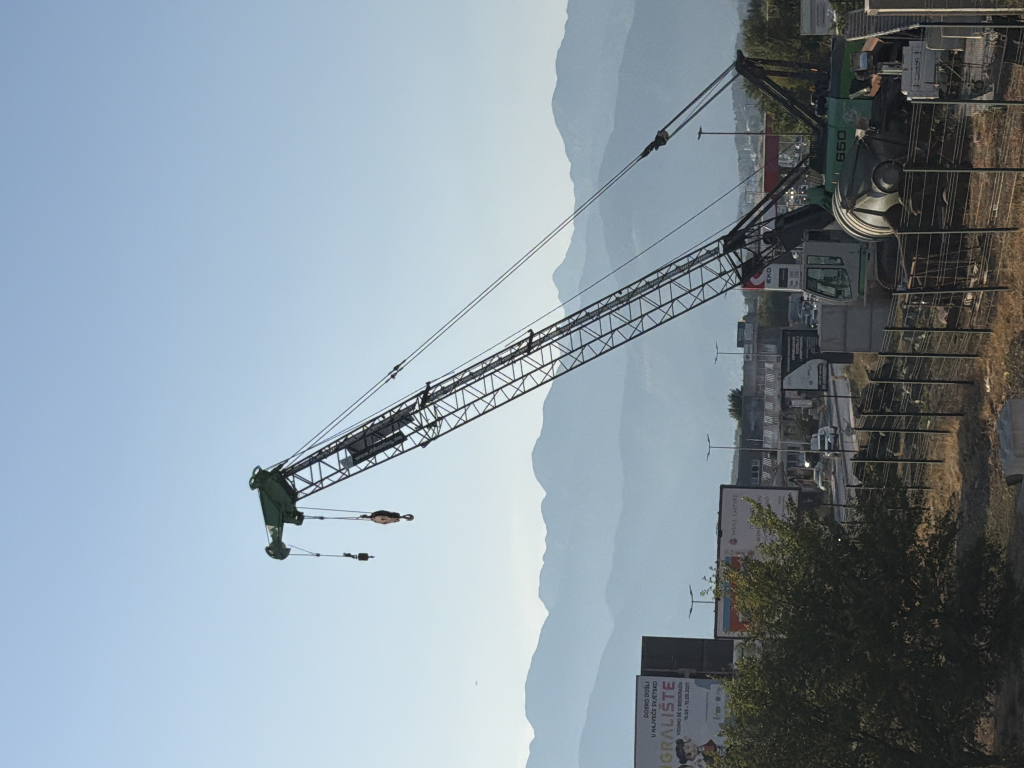 The image depicts a construction site under a clear blue sky, with a large green crane dominating the scene. The crane's boom extends diagonally across the frame, with its base obscured by other construction equipment. In the background, a range of hazy blue mountains forms a layered silhouette against the sky. Below the mountains, several billboards are visible, one prominently displaying text in what appears to be Serbian or a similar Slavic language. A line of trees and shrubs partially obscures the view of the road and buildings in the middle ground, where several cars and a truck can be seen. The foreground is a mix of dry grass, a metal fence, and concrete barriers. On the crane itself, the number "650" is visible in green lettering. The overall impression is one of ongoing construction in a semi-urban area with a mountainous backdrop.