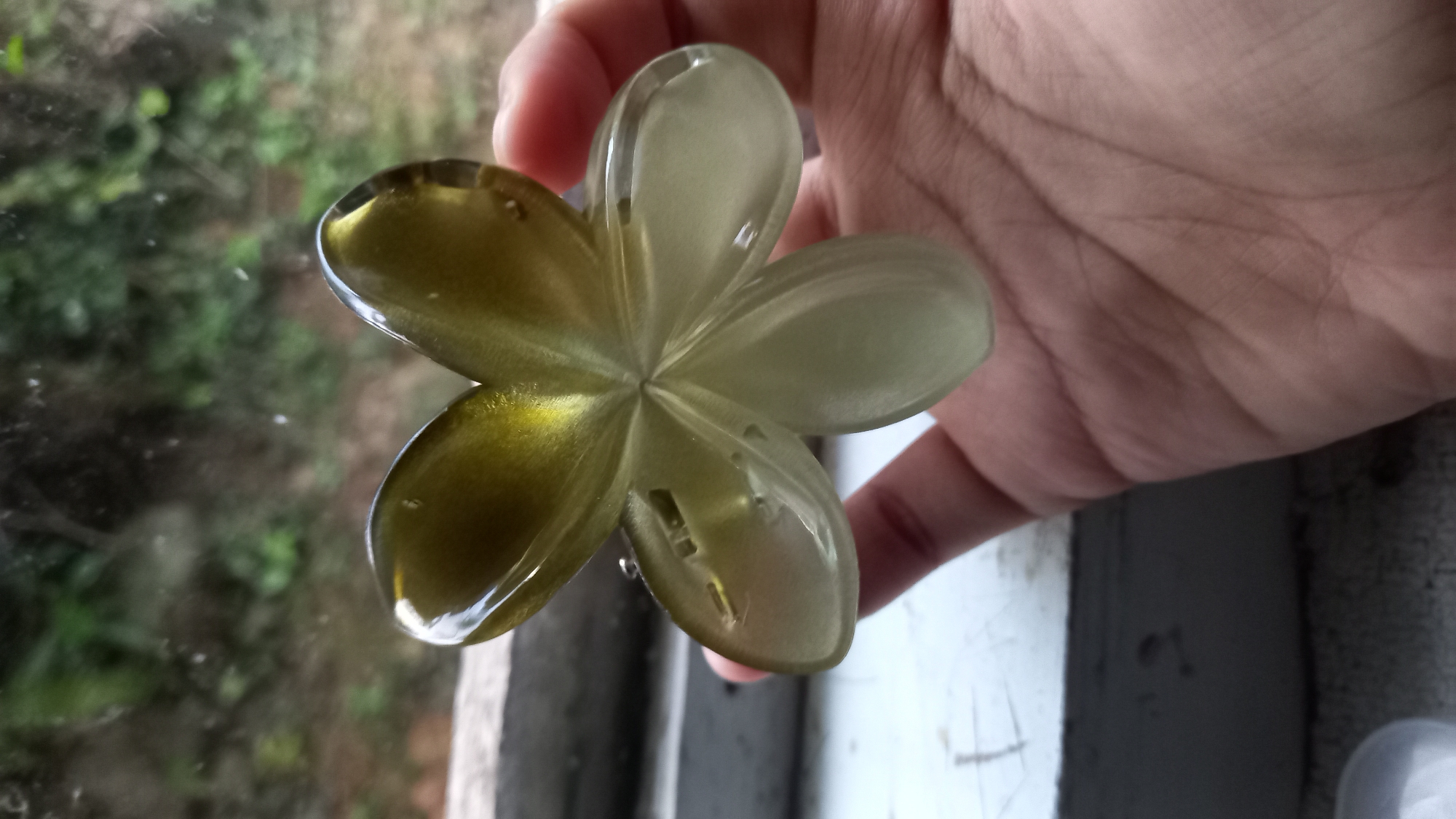 A person's hand is holding a translucent, flower-shaped object. The object is predominantly light green with hints of yellow. The background suggests an outdoor environment, possibly a window looking out onto foliage and trees. The scene is captured in Leuwisadeng, Indonesia.