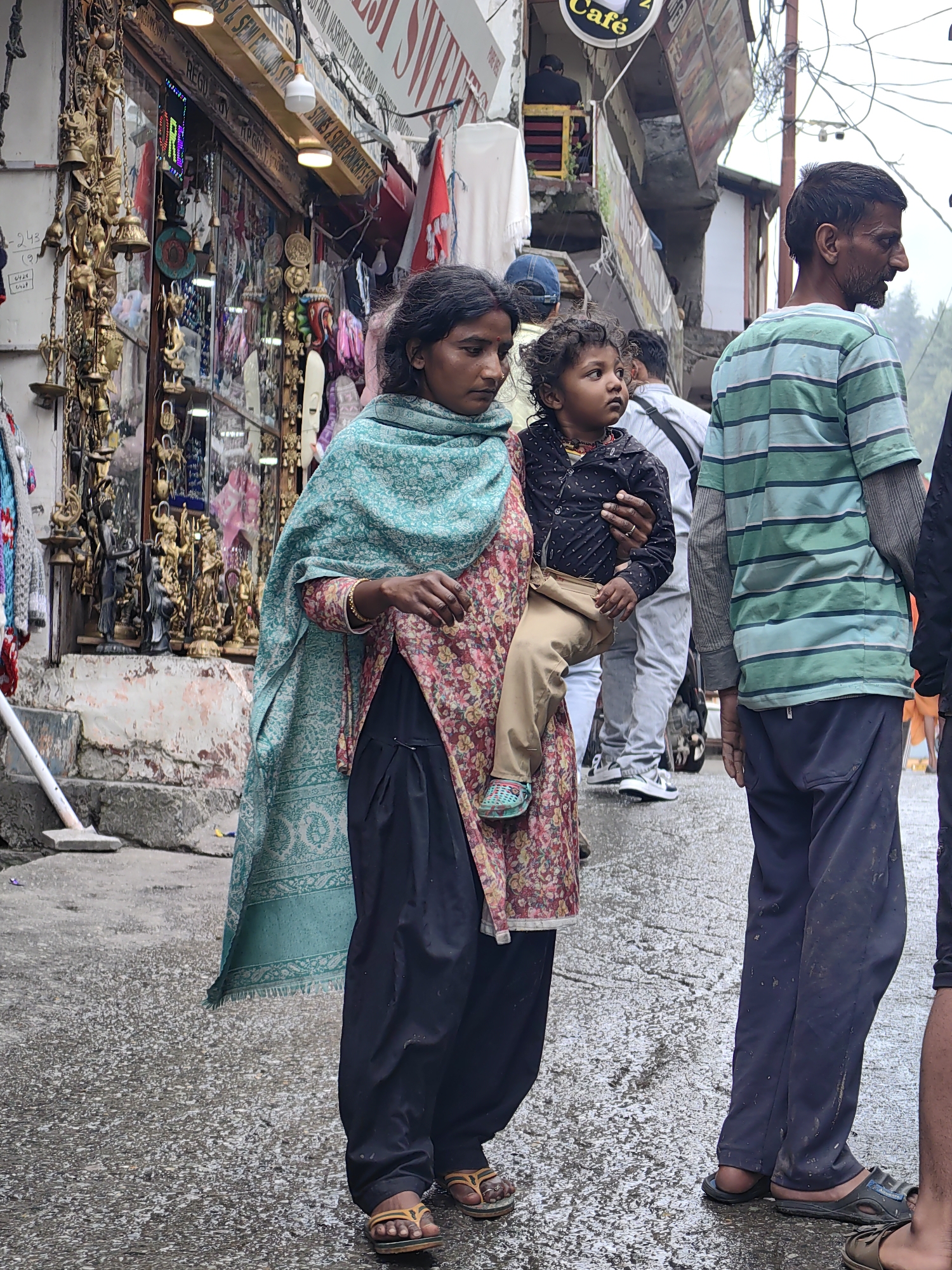In Vashisht, India, a woman carries a young child in her arms while standing on a wet street in front of a market stall. The woman is wearing a floral print dress and a teal scarf. The child is dressed in a black shirt and tan pants. Behind them, a man in a green and blue striped shirt stands on the street. The market stall displays various items, including statues and bells. The street is wet and reflective, indicating recent rain. The overall scene depicts a typical street view in a busy Indian town.