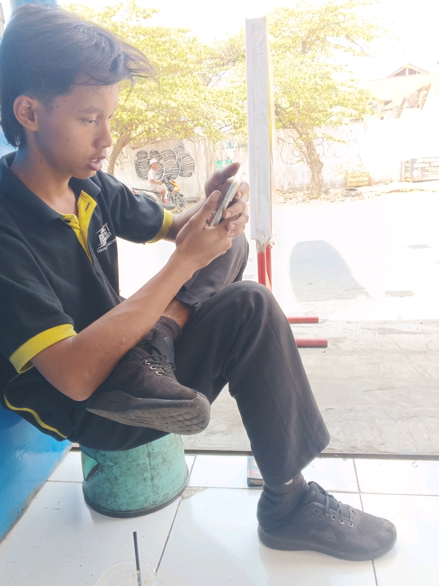 A young person, likely male, with dark, styled hair, is seated outdoors, holding a mobile phone and looking at it. They are wearing a black polo shirt with yellow trim and black pants. Their right foot rests on a light blue, weathered plastic bucket. Their left foot is also visible, wearing a black athletic shoe. In the background, there is a wall with graffiti, some greenery, and a building. A motorbike with a rider is also visible in the distance. The scene suggests a casual setting, possibly on a street or in a public area, in Taman, Indonesia. The lighting is bright, indicating daytime.