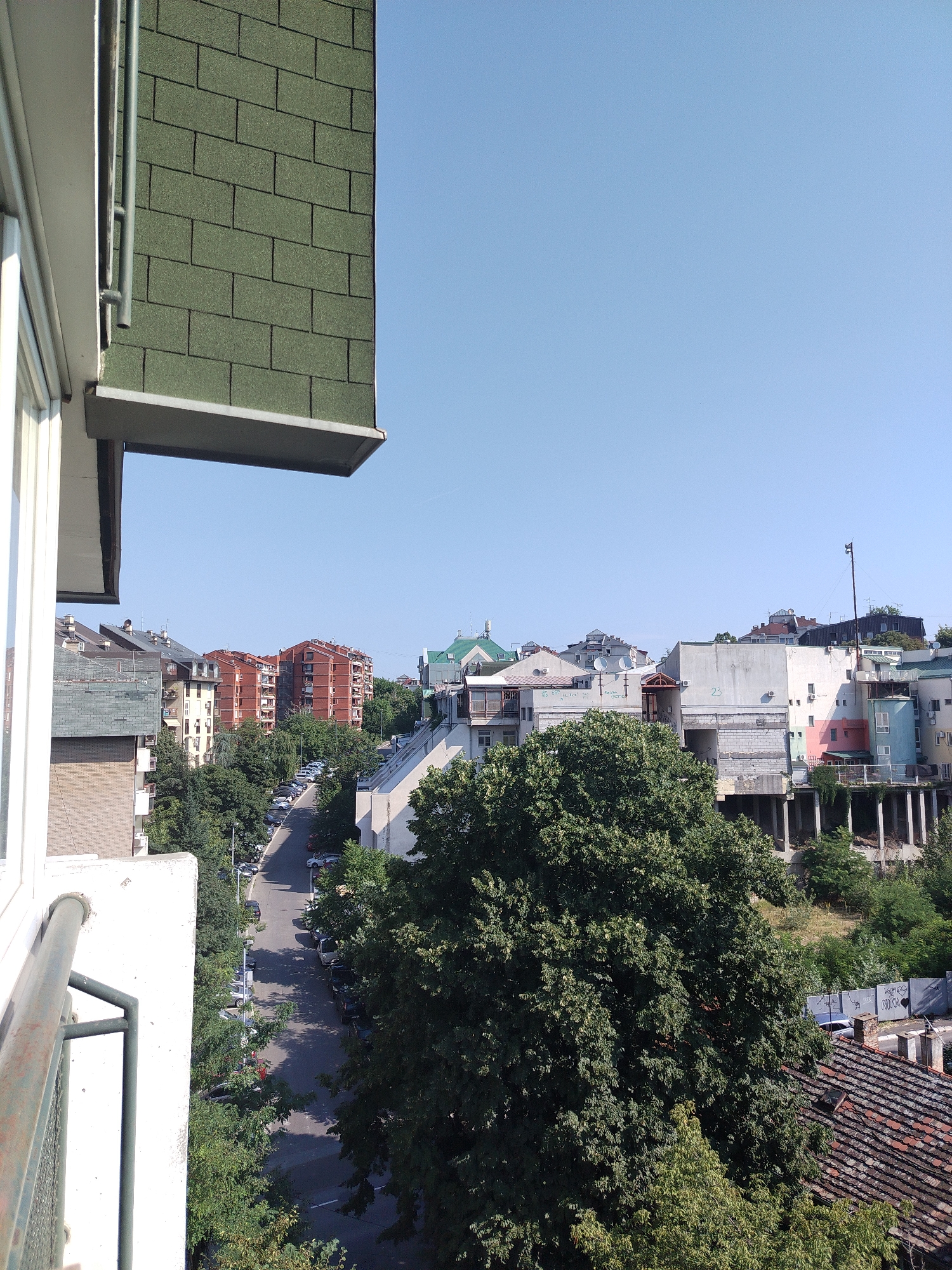 This is an outdoor, eye-level, medium-shot photograph taken on a sunny day, likely in the Cukarica Urban Municipality of Serbia, as indicated by an external source. The perspective is from a balcony, looking down and across a street lined with parked cars and trees.
In the foreground, the edge of a white building with a metal railing is visible on the left. Above this, a section of a dark green, shingled roof juts out from another building.
The midground is dominated by a winding asphalt street with cars parked along both sides, indicating a residential or mixed-use urban area. Mature green trees line the street, casting shadows on the road. Across the street, a variety of buildings are visible, ranging from multi-story brick apartment buildings with red-tiled roofs to lighter-colored structures, some with visible balconies and architectural details. One prominent building in the mid-distance is made of red brick and appears to be an apartment complex.
In the background, a collection of buildings, including some with flat roofs and others with pitched roofs, are spread across a gentle slope. The sky is clear and blue, suggesting good weather conditions. The overall impression is of a moderately dense urban environment with a significant presence of greenery. There are no people or animals clearly visible in the frame.