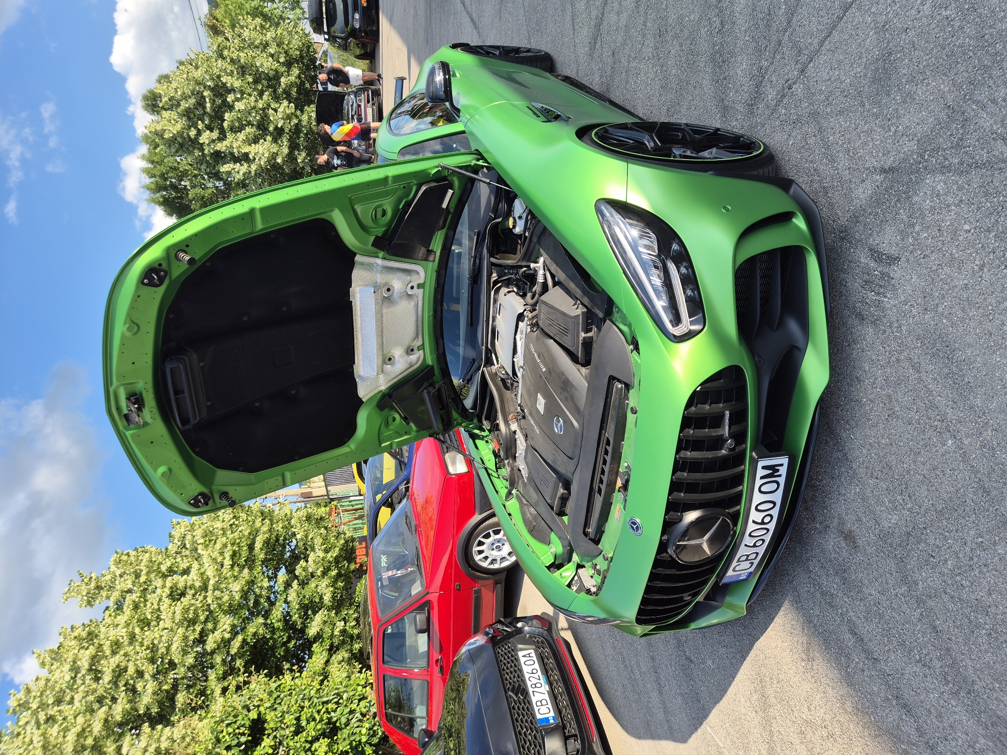 The image depicts an outdoor car gathering under a bright, partly cloudy sky. In the foreground, a matte-lime-green Mercedes-AMG GT R (or similar GT variant) is parked, angled towards the left, with its hood open, revealing the engine bay. The front license plate reads "CB 6060 OM" and includes a Bulgarian country code identifier. The vehicle features black multi-spoke wheels with a green accent ring.
To the left of the Mercedes-AMG, a black car, possibly a modern performance hatchback, is partially visible, showing its grille with red lower trim and a Bulgarian license plate "CB 7826 OA." Behind it, a red two-door Volkswagen Golf Mk2 with white multi-spoke wheels is parked. Further in the background, a yellow car is partially obscured. On the right side of the background, a black Volkswagen Golf Mk4 equipped with a roof rack and roof box is visible, also with its hood open.
Three men are positioned in the background near the black Golf Mk4. One wears a black t-shirt, another a multi-colored striped t-shirt, and the third a white t-shirt and white shorts. They are observing the scene. The ground is paved with grey asphalt or concrete, showing strong shadows from the sunlight originating from the left. The background is dominated by lush green trees with some lighter-colored foliage under a blue sky with scattered white clouds. The location is an unspecified outdoor venue in the city of Unknown, Unknown, with multiple vehicles displaying Bulgarian license plates.