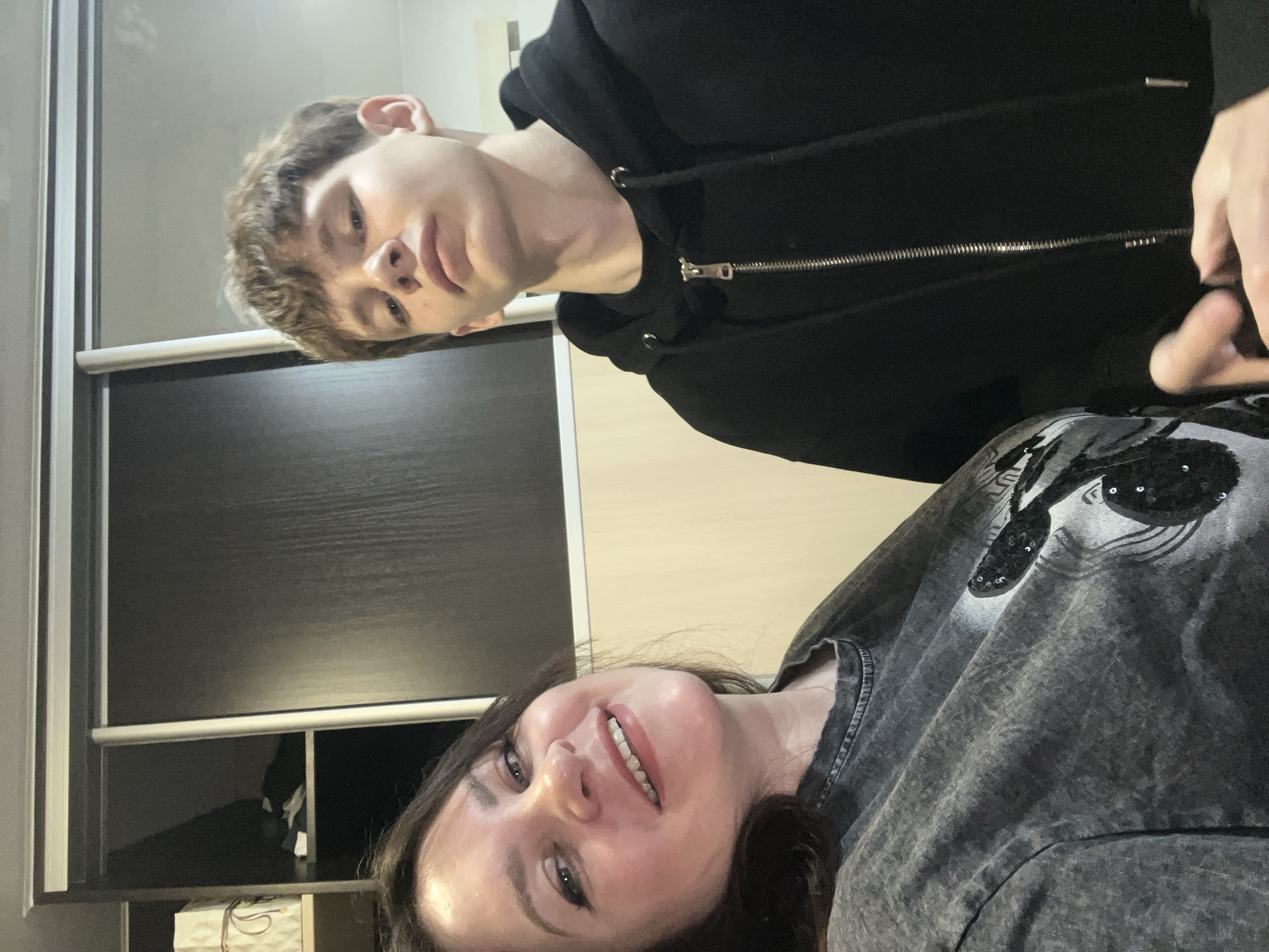Two individuals are present in an indoor setting. A young man, dressed in a black hooded sweatshirt, is positioned in the upper portion of the image. He has light skin and brown hair. Below him is a woman with dark hair, wearing a gray t-shirt. She is smiling. The background includes what appears to be a cabinet or wardrobe, and partially visible, a window. It is not possible to confirm the location in Noviy, Russia based on the provided image.