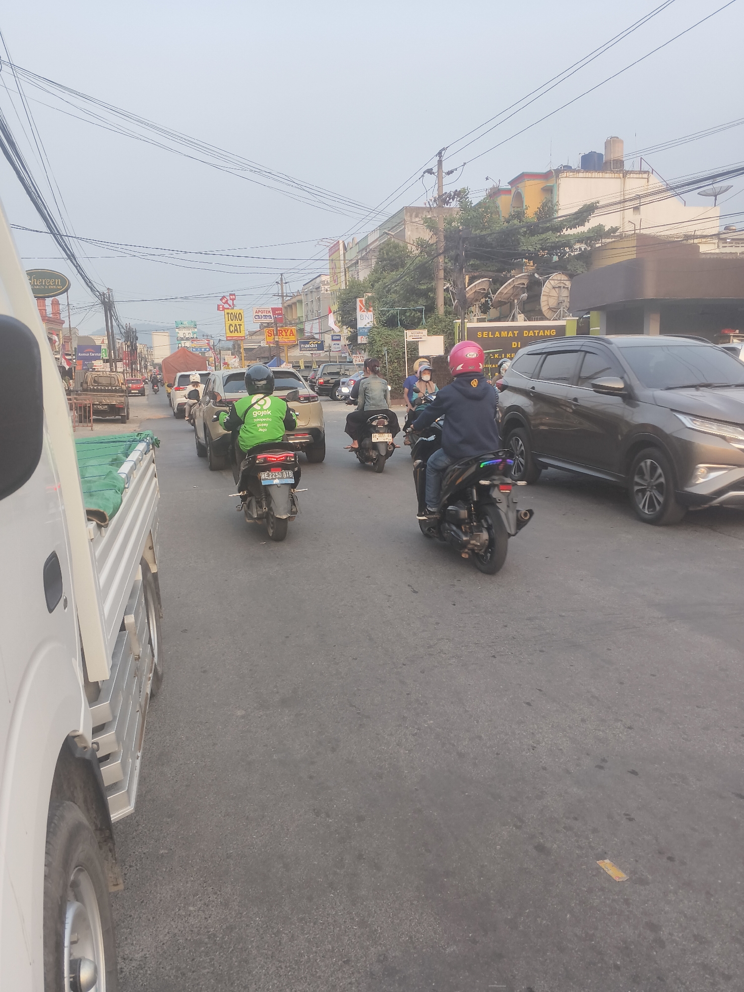 In Bandar Lampung, Indonesia, a street scene unfolds with multiple vehicles and pedestrians. The foreground is dominated by the side of a white truck, its silver metallic cargo bed and wheel visible. To the right, the asphalt road shows signs of wear and tear.
Several motorcycles and cars are present. A motorcycle with a green jacketed rider, bearing the "Gojek" logo, is moving forward. Another motorcycle is carrying two individuals, one in a blue jacket and pink helmet, the other in a dark jacket. A dark grey SUV is parked to the right of the motorcycle. Further down the road, more cars and motorcycles are visible, suggesting moderate traffic.
The background reveals a row of buildings with various signs, including "Shereen," "Toko Cat," and "Apoter Cantiya." Overhead, a dense network of power lines crisscrosses the sky, obscuring the view of the sky. The atmosphere appears hazy, possibly due to pollution or the time of day, with a bright sky suggesting daylight. The overall impression is a busy urban environment.