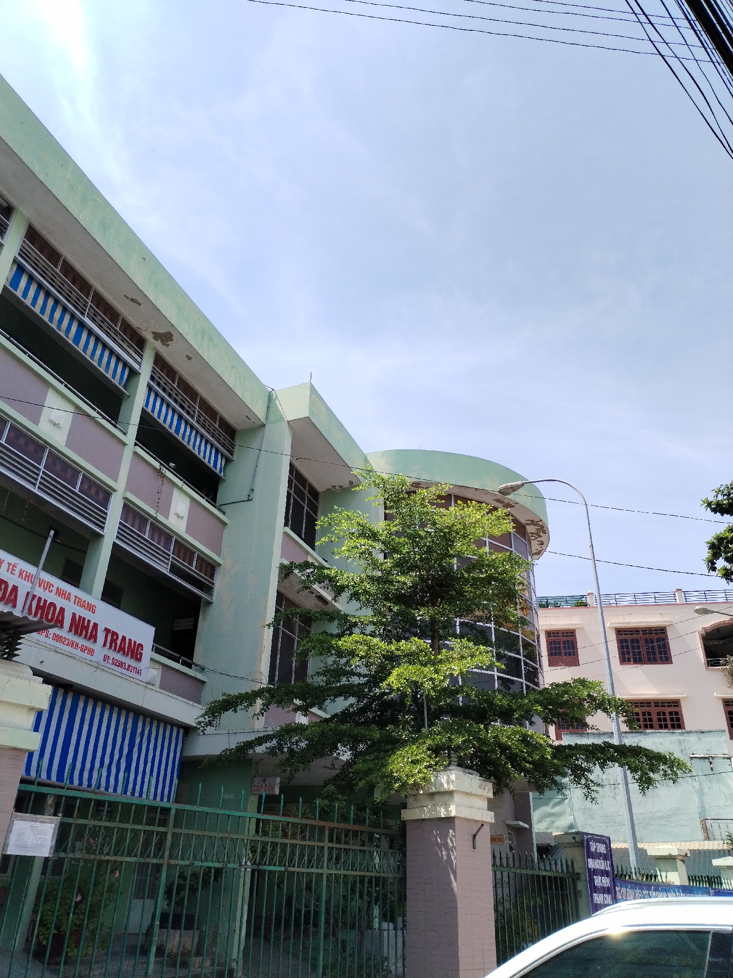 The image shows a multi-story building in a city, likely Nha Trang, Vietnam, given the signage. The building has a pale green facade with several rows of windows. Some windows are covered with blue and white striped awnings. A large tree with green foliage is growing in front of a curved section of the building. Electrical wires are visible against a clear sky. A green metal fence runs along the front of the property, and a sign with Vietnamese text is attached to the fence. Another sign, also with Vietnamese text, is visible on the building's facade. The overall scene appears to be an exterior view of a medical facility or similar institution. There are no people or vehicles prominently featured in the image.