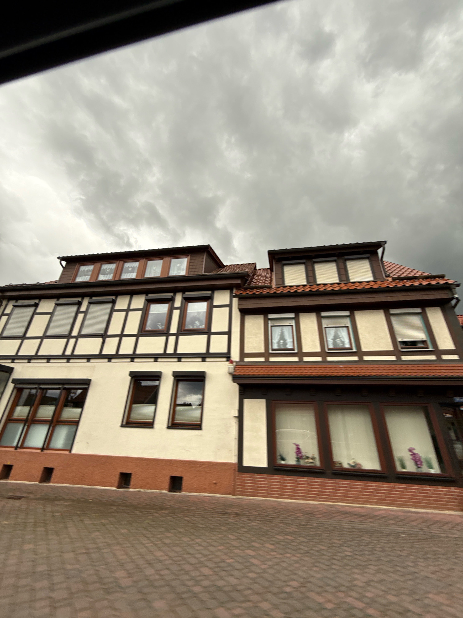 The image shows a two-story building with a dormer roof, characteristic of German Fachwerk architecture. The building has a cream-colored facade with dark brown timber framing. The roof is tiled with reddish-brown tiles. The lower story features large, dark-framed windows, some with reflective surfaces showing the cloudy sky. The upper story has smaller windows, many equipped with roller shutters. There are small, dark rectangular openings along the base of the building on the lower story. The ground in front of the building is paved with reddish-brown cobblestones. The sky is heavily overcast with dark, dramatic clouds. This building is located on Ravensbergstrasse in Bad Sachsa, Germany.