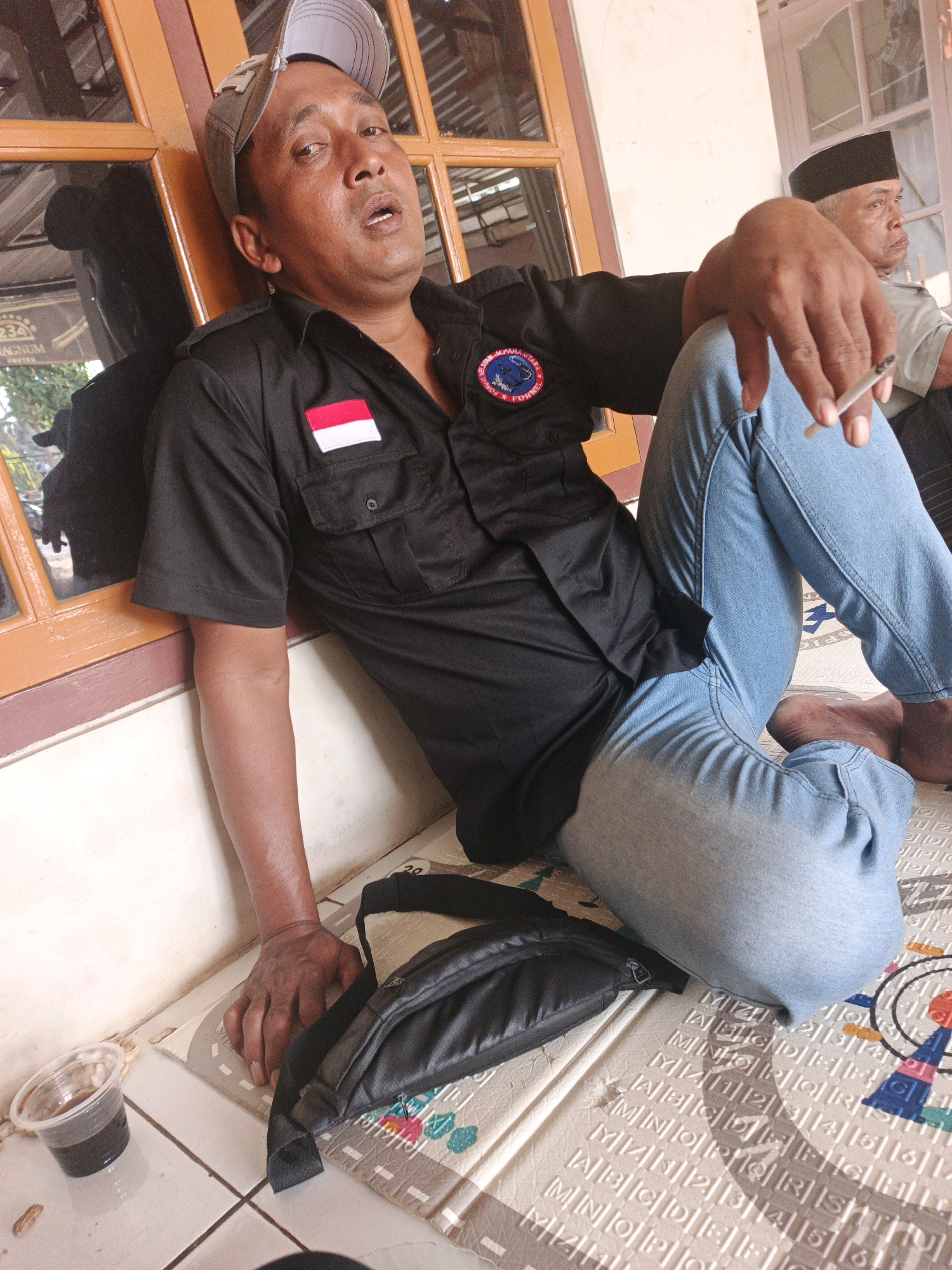 A man, appearing to be of Indonesian ethnicity, is shown resting against a window frame. He is wearing a black short-sleeved shirt with a red and white flag patch on the left chest and a circular emblem on the right. Light blue denim jeans are worn. His eyes are closed or looking down, and his mouth is slightly open. He is holding a lit cigarette in his right hand, which is resting on his left knee. Next to him, a black fanny pack is placed on a mat. A small clear plastic cup containing a dark liquid is on the floor to his left. In the background, another man wearing a black cap and a light-colored shirt is visible. The setting appears to be outdoors or in a semi-enclosed space with wooden window frames and a tiled floor. The overall impression is of a casual and possibly tired moment. The location is specified as the city of Kaliaman, Indonesia.