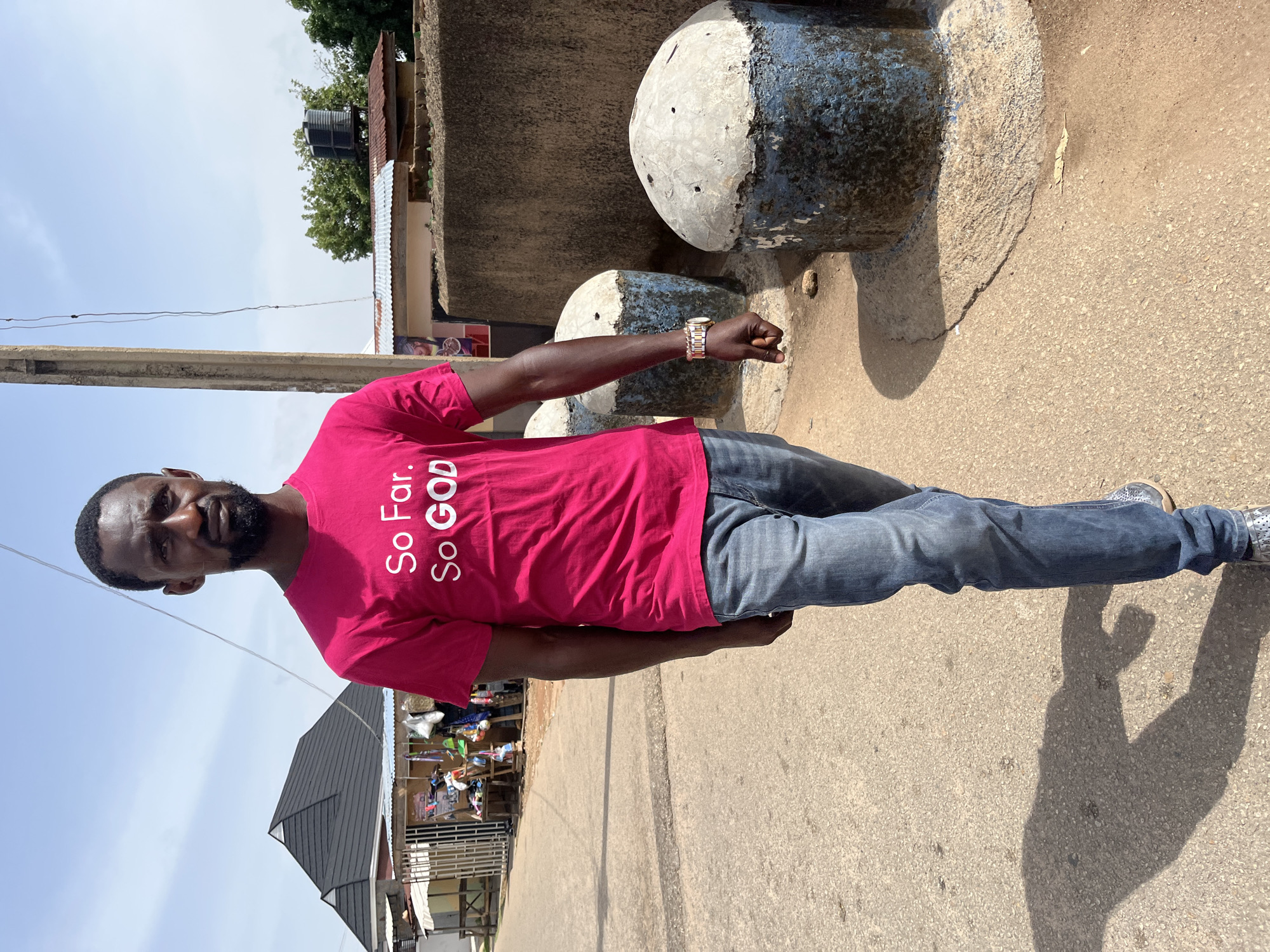 A man stands on a paved road near what appears to be the city of Jos, Nigeria. The man wears a pink t-shirt that reads "So Far. So GOD." He is wearing blue jeans and light-colored shoes. He also has on a gold wristwatch. He is standing near a short cement structure with two round concrete objects on top, painted with a dark blue and white pattern. Behind him, there's a building with a dark gray roof and shops set up beside the road. A utility pole is located to the left of the man. The sky is light blue.