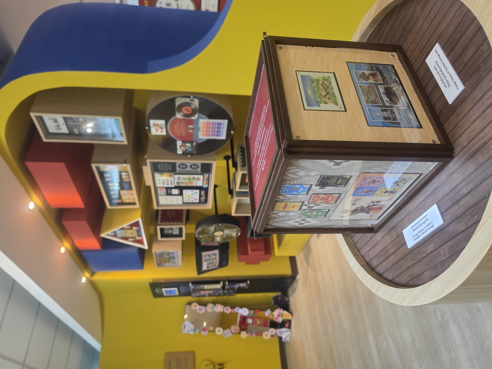 The image depicts an interior scene, potentially a museum or exhibition space, filled with colorful, modern displays. Dominant hues are yellow and blue, which cover walls and ceilings. The room features a variety of showcases and displays, some containing stamps and other artifacts. One display features what appears to be stamps. A large wooden circular pedestal supports a framed exhibit of stamps, and signs provide information in both Thai and English, specifying "Postage Stamps of UNESCO Intangible Cultural Heritage" and "Postage Stamps of Thai World Heritage Sites".
A mirror adorned with decorative items and other objects on shelves and displays indicates the space is a curated exhibit. Given the presence of Thai text and the references to Thai World Heritage Sites, the location is likely within Bangkok, Thailand.