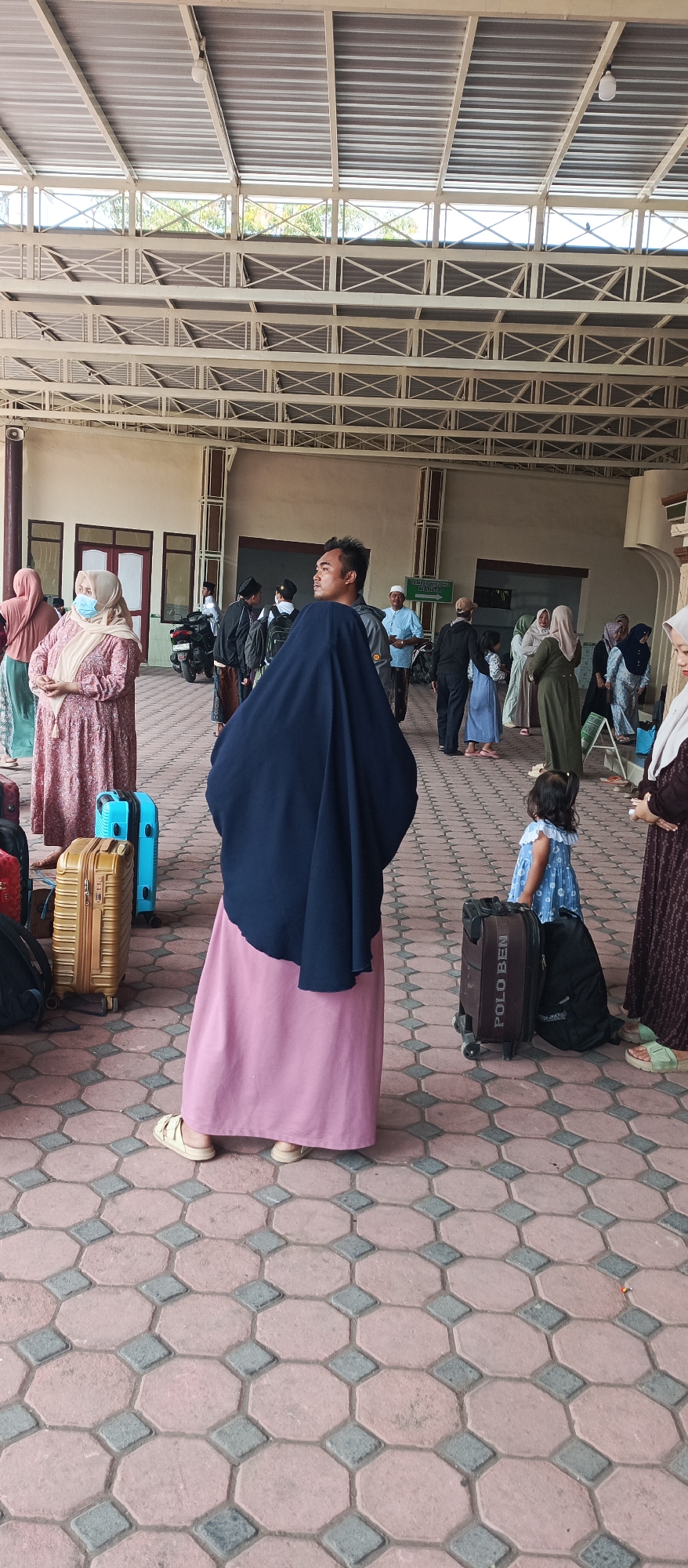The image depicts a gathering of people, likely in a public waiting area or entrance of a building in Kapongan, Indonesia. The foreground is dominated by the back of a person wearing a dark blue head covering and a long pink skirt, with light-colored sandals. To the left of this person are two suitcases, one gold and one blue, and a black bag. A young child in a blue dress stands near a black suitcase with "POLO BEN" visible on it. In the background, a group of individuals, some wearing traditional Islamic attire, are visible. One man with short hair is looking to the left. A motorcycle is parked in the middle ground. The environment features an open-air structure with a metal roof supported by beams and columns, and a tiled floor with an octagonal and diamond pattern. Signs are visible on the walls of the building. The overall scene suggests a travel or waiting scenario.