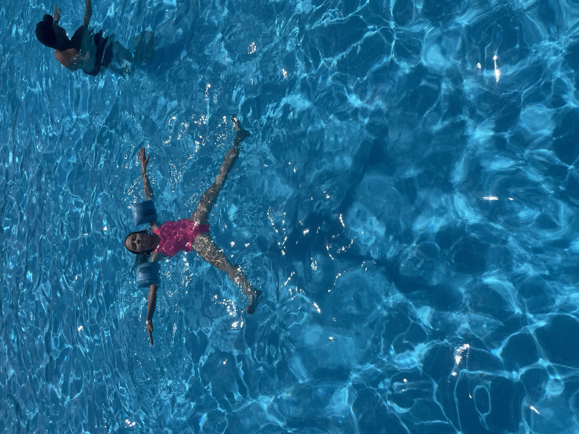The image captures an overhead view of two people swimming in a bright blue swimming pool. One individual, a child, is floating on their back, wearing a pink swimsuit and blue arm floaties. Their arms are outstretched, and their face is tilted upwards, suggesting a relaxed state. The second individual, an adult, is visible in the upper left portion of the image, partially submerged and appearing to be swimming or treading water. The water is illuminated by sunlight, creating shimmering patterns of light and shadow on the surface, indicating it is daytime. The setting is a swimming pool, likely outdoors given the natural light. The scene evokes a sense of leisure and enjoyment, likely during warm weather. No visible text is present in the image. The location context is Пристанное, Russia.