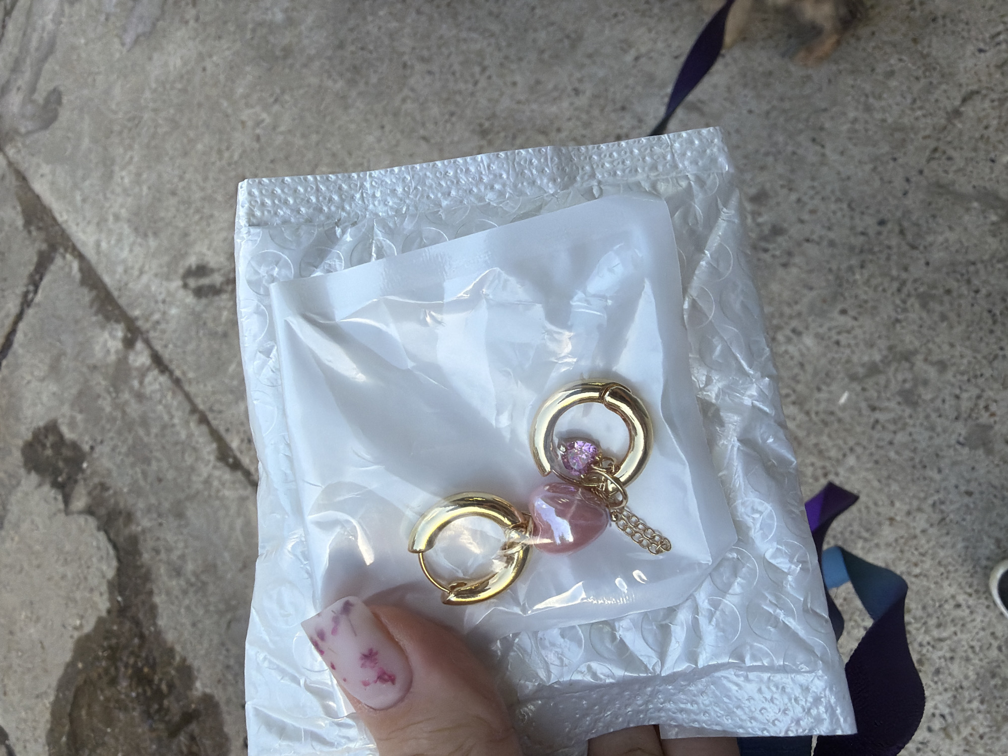 The image shows a person holding a package containing a pair of earrings. The earrings are gold with pink heart-shaped charms and a small pink gem. The package is made of clear plastic with bubble wrap inside. The background is a grey concrete surface. A small dog is visible in the upper right corner of the image, and a piece of purple ribbon is also present. The lighting suggests it is daytime, and the weather appears to be mild. The image is taken from a top-down perspective, focusing on the earrings and the hand holding the package. There is no visible text in the image. The scene is casual and appears to be outdoors.
