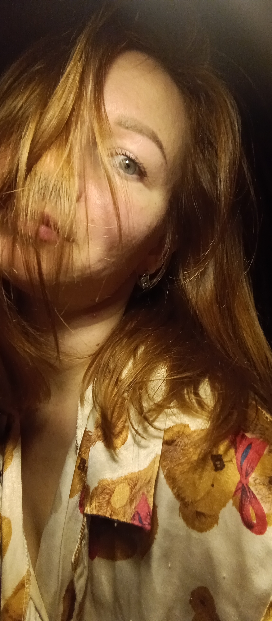A medium shot shows a woman with auburn hair, part of which is covering her face and resembling a mustache. Her left eye is visible and appears to be grey. She is wearing an earring on her left ear. The woman is wearing a pajama top or robe with a teddy bear print, one of which has a red and blue bow. The background is dark and out of focus. Location: Unknown, Unknown.