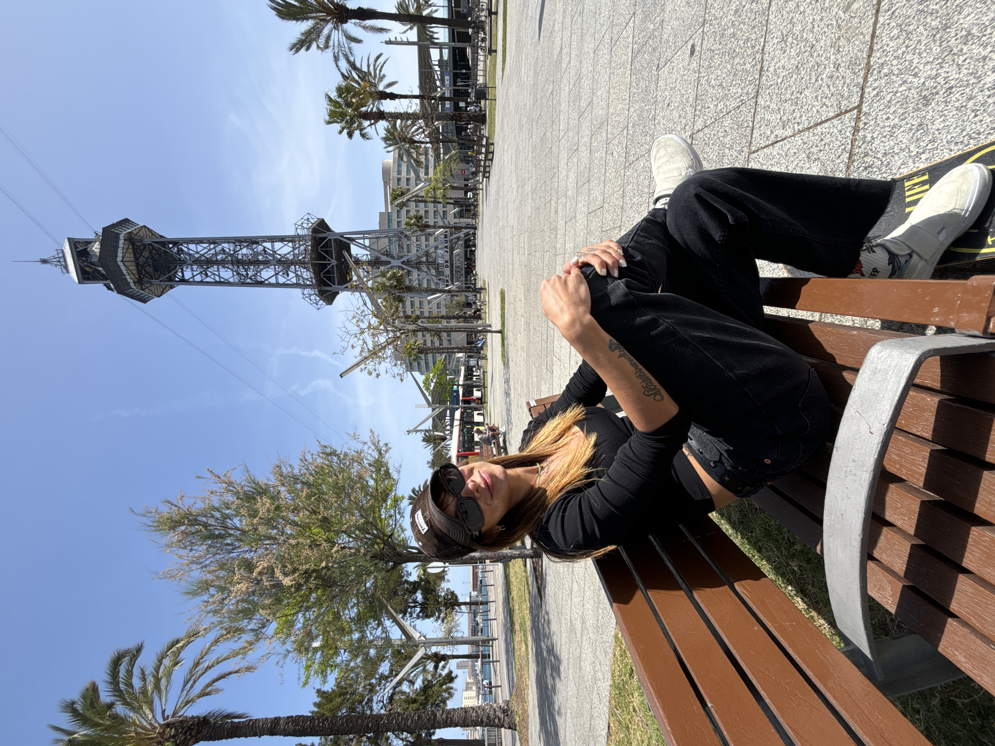 The image captures a young woman relaxing on a brown wooden park bench on a sunny day in Barcelona, Spain. She is dressed in a black long-sleeved top, black pants, and light-colored sneakers, with a skateboard placed under her feet. She wears sunglasses and a dark baseball cap with a white logo, partially visible as "VANS." Her hands are clasped over her bent knees, and a script tattoo is visible on her left forearm.
The scene is an outdoor urban environment, likely a promenade or park area. The ground is paved with light-colored square tiles, and strips of green grass run alongside. Several tall palm trees and other leafy trees are scattered throughout the background. A prominent landmark, the "Torre de Jaume I" (Jaume I Tower), part of the Port Cable Car system, stands tall against the bright blue sky. Other buildings are visible in the distance. The weather is clear and sunny, indicated by the bright sky and distinct shadows. The woman appears relaxed, looking towards the camera with a subtle smile. Visible text includes "TORRE DE JAUME I" on the tower and "LIFE" on the skateboard.