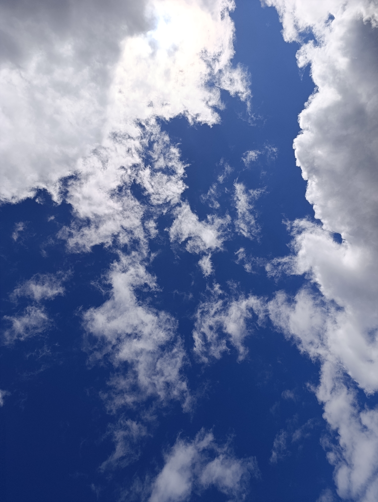 The image shows a view of the sky. The sky is predominantly blue, with scattered cirrus clouds and cumulus clouds. The clouds are white and appear fluffy. The sun is partially obscured by clouds in the top portion of the photo. The photograph was likely taken during the daytime. The location in the city of Chaikovsky, Russia, is only inferred information.