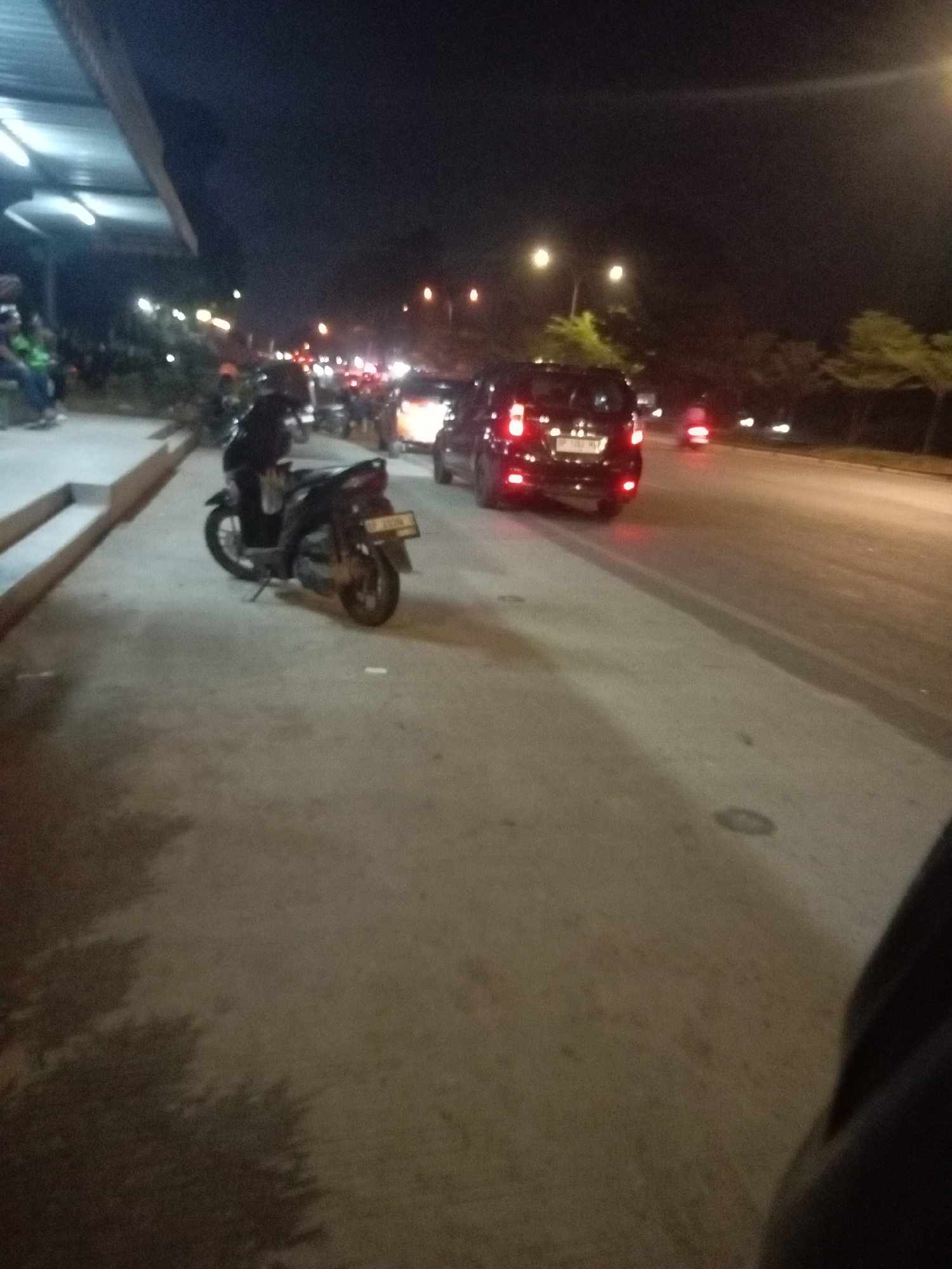 A nighttime street scene in Batam, Indonesia, captures traffic and pedestrians. A dark-colored motorcycle is parked on the sidewalk, angled towards the street. Behind it, several cars are visible, some with their brake lights illuminated, indicating slow-moving or stopped traffic. The road appears to be a multi-lane street with streetlights casting a warm glow. On the left side of the image, a covered walkway or building structure is present, with some people seated underneath. Trees with sparse foliage line the right side of the road, silhouetted against the dark sky. The overall atmosphere is one of a moderately busy urban environment at night.