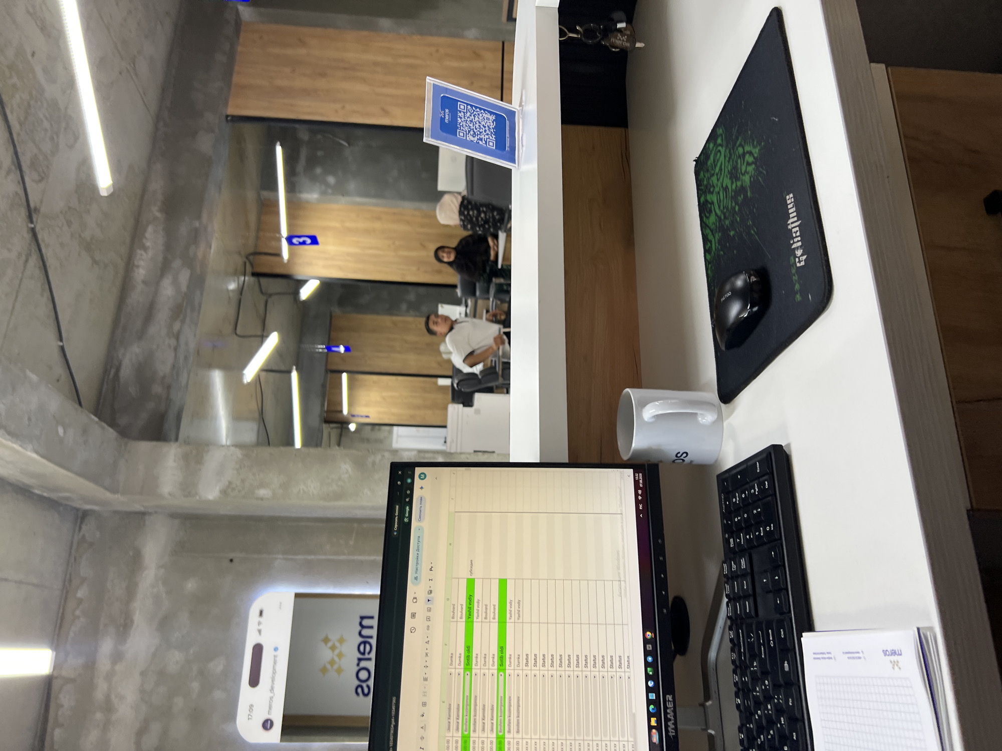 The image depicts an office environment in Shahrixon, Uzbekistan. In the foreground, a desk is laden with office supplies, including a computer monitor displaying a spreadsheet, a keyboard, a mousepad with a gaming mouse, a white mug, and a notebook. The monitor's screen shows a spreadsheet with columns labeled "00:00," "Status," "Status," and "Status," suggesting data entry or tracking. The computer's taskbar indicates the time as "13:32" on "07.08.2020," and displays the operating system as "Windows." In the background, the office space features concrete walls and partitions with wooden accents. Several individuals are visible, including two women and one man, seated at desks or standing. One woman is wearing a hijab and an abaya, while the other wears a hijab and a patterned dress. The man is dressed in a white polo shirt and dark trousers. Blue signs with numbers are mounted on the partitions, indicating office numbers or zones. A prominent display on the wall shows a mobile phone interface with the "meros_development" logo. A QR code is displayed on a stand on the counter in the mid-ground, likely for check-ins or information access. The overall impression is that of a functional workspace with a modern aesthetic.