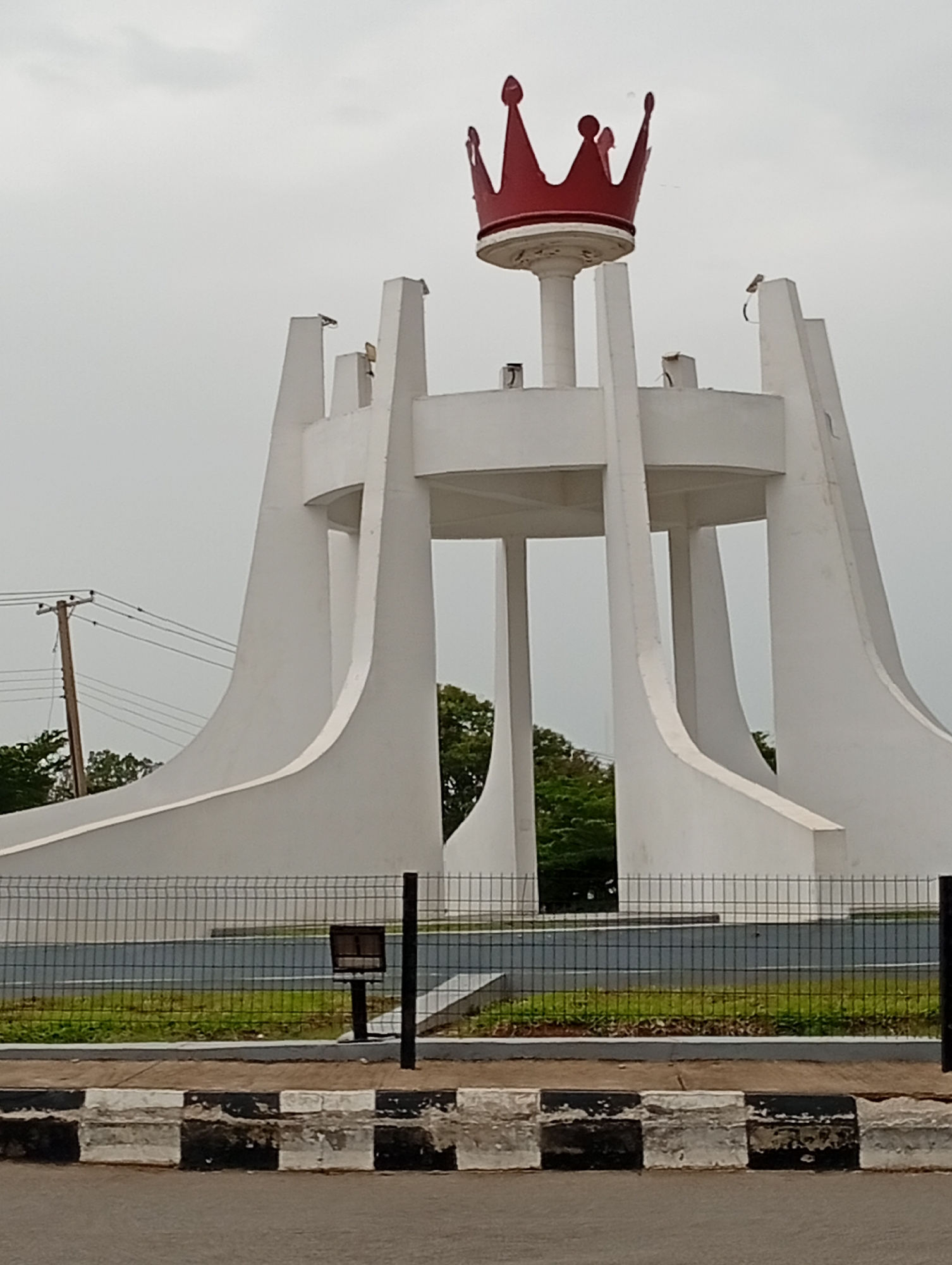 The image shows a monument in the city of Jos, Nigeria. The monument is white and has a red crown on top. It is surrounded by a metal fence and grass. There are trees and utility poles in the background. The sky is overcast. The monument appears to be a landmark or a tourist attraction. The scene is captured from a slightly low angle, looking up at the monument.