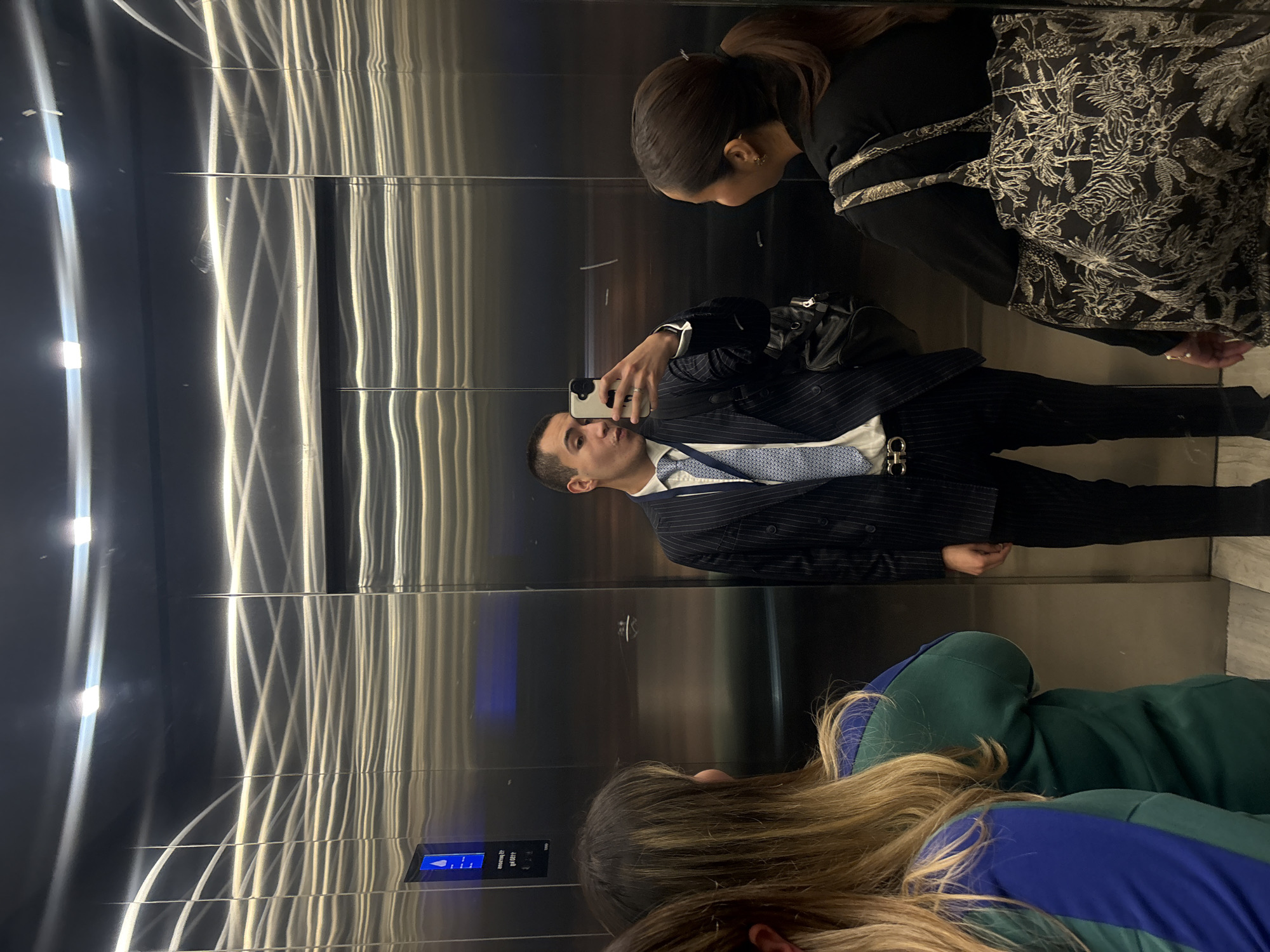 Three individuals are in an elevator. One man, wearing a pinstripe suit, stands with his arm raised, holding a smartphone, apparently taking a selfie in the elevator's mirrored interior. Above and to the right of him, a woman with her hair in a ponytail looks down, partly obscured. Below him, another woman with long blonde hair, wearing a green and blue outfit, is visible from the back of her head and shoulders. The elevator's walls are metal. The location is San Bernardino Tlaxcalancingo, Mexico.