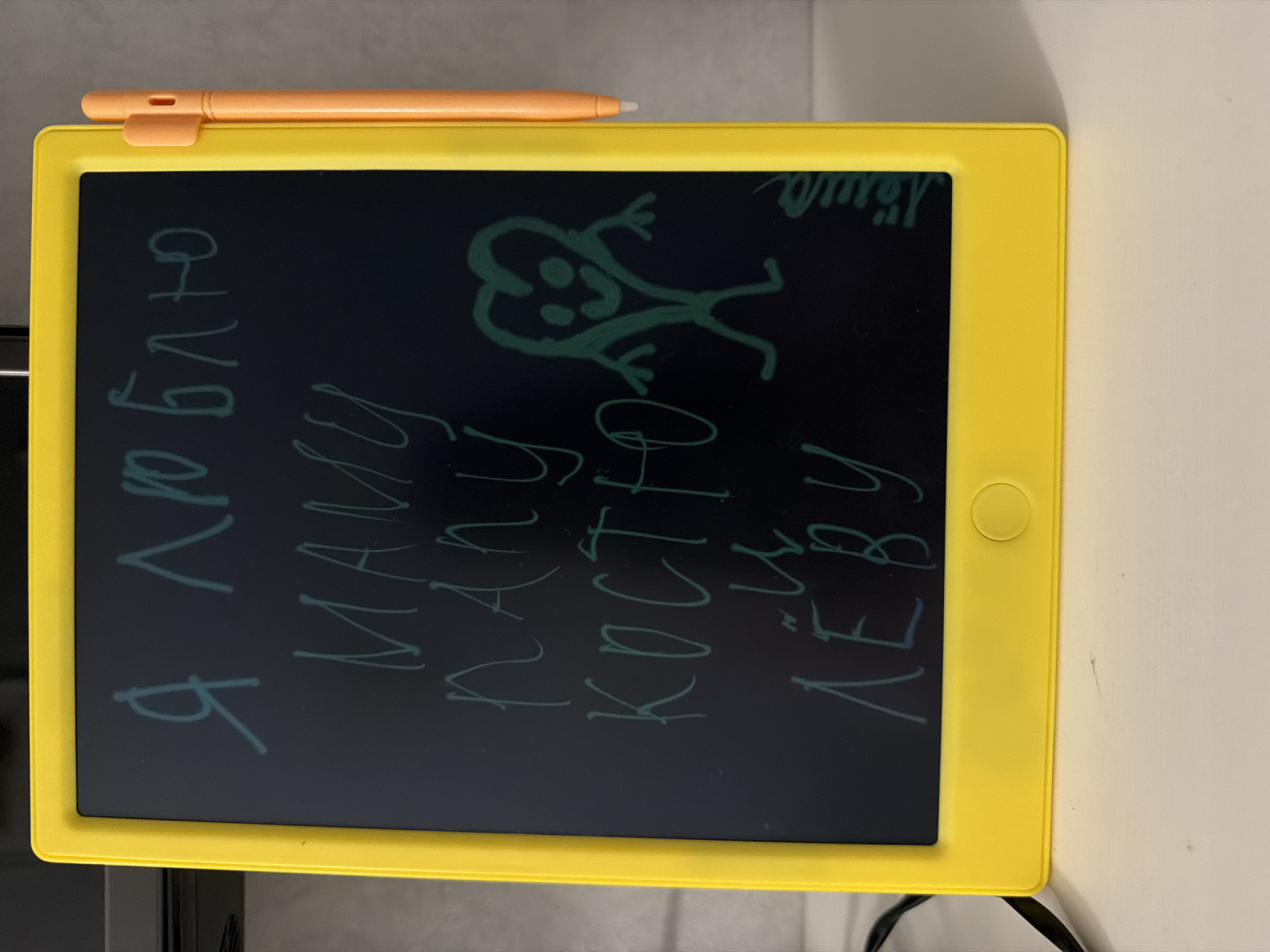A yellow LCD writing tablet is on a surface. The screen displays a drawing of a stick figure and text in Cyrillic: "Я люблю Маму Нашу Костю Леву" and "Шмяу". An orange stylus is attached to the top of the tablet. The background shows a neutral colored wall and a portion of what appears to be a dark appliance. The location is not specified as being in Votkinsk, Russia and no people, actions, or interactions are observed.
