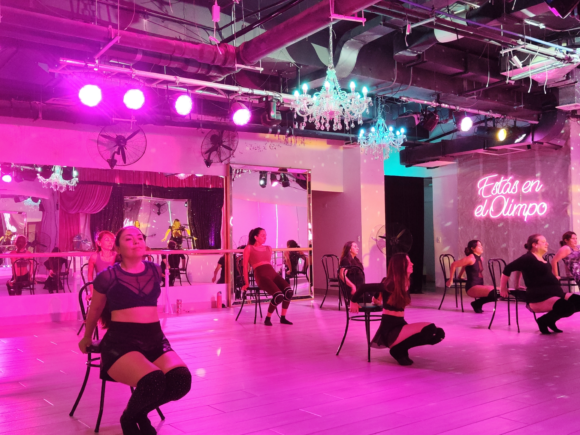 This image depicts a dance class or performance taking place in a studio with pink lighting. The room features a wooden floor, a mirrored wall on the left, and a neon sign on the right wall that reads "Estás en el Olimpo" in a cursive font. Several women are present, dressed in various dance attire, including leotards, shorts, and thigh-high boots. Many of them are positioned on or around chairs, engaging in dance movements. The overall ambiance is energetic and theatrical, enhanced by the vibrant lighting and the studio's decor. It is highly probable that this studio is located in Santiago, Chile, given the Spanish text on the neon sign.