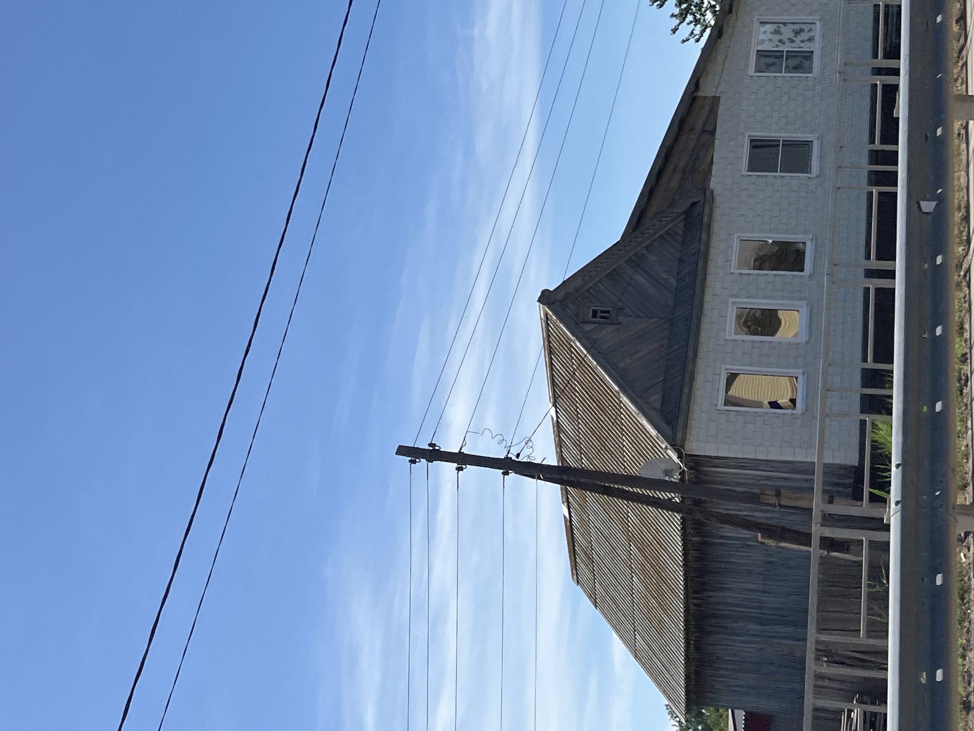The image depicts a rural scene under a clear blue sky with wispy clouds. In the foreground, a grey metal guardrail runs horizontally across the bottom of the frame. Behind the guardrail, a weathered wooden fence encloses a property. The main subject is a two-story house with a corrugated metal roof. The left side of the house appears to be clad in vertical wooden planks, while the right side is constructed of light-colored brickwork. Several windows are visible on the brick section of the house, some with curtains. A wooden utility pole with multiple wires attached stands prominently to the left of the house, with a satellite dish mounted on its upper section. The overall impression is that of a somewhat aged but functional dwelling in a non-urban setting.
