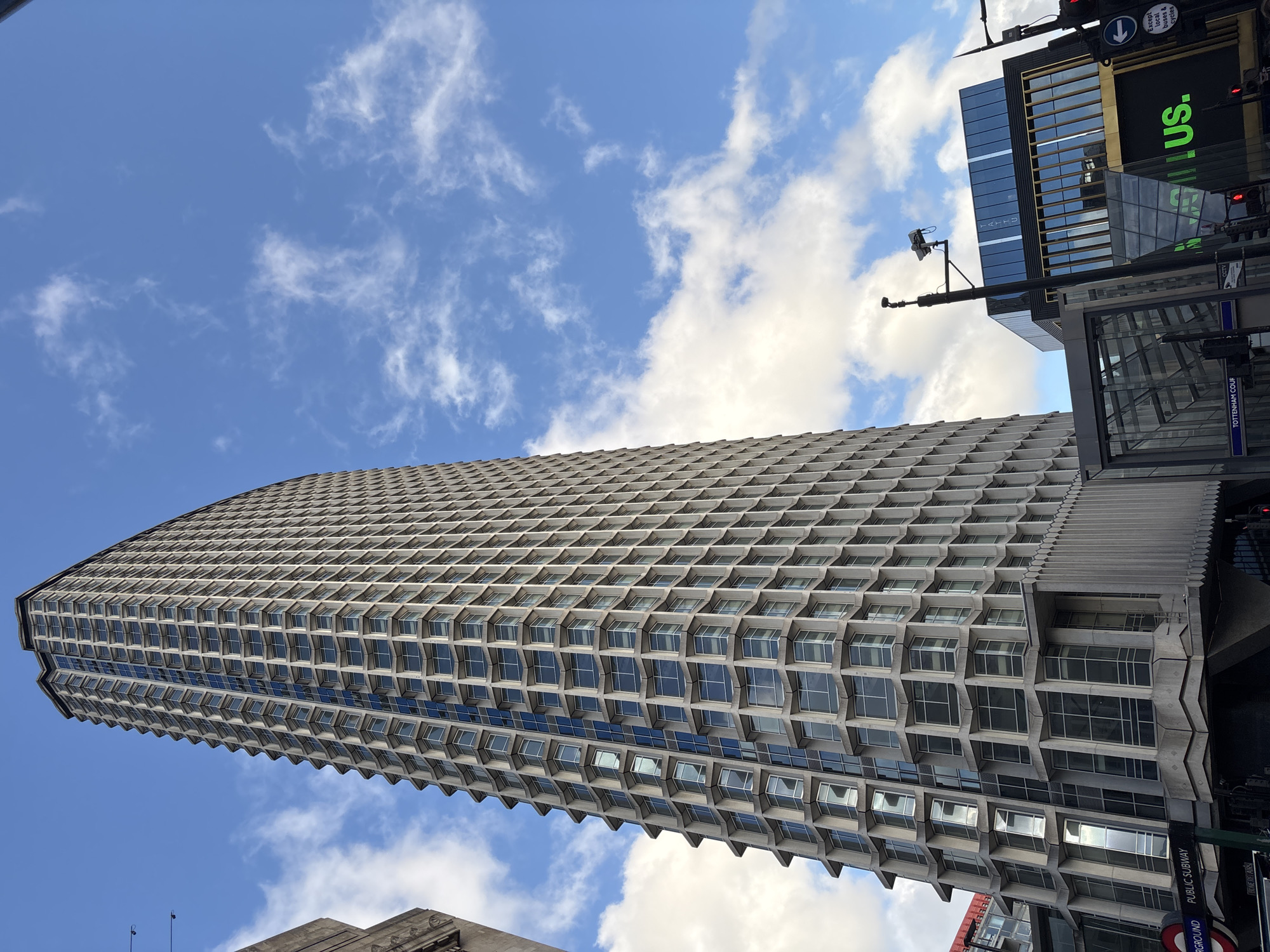 The image shows a tall, modern building in London, United Kingdom, angled upward against a blue sky with scattered clouds. The building has a distinctive honeycomb-like facade. A sign for Tottenham Court Road Underground station is partially visible in the lower right, suggesting the building's location. A partial billboard with the text "US." is seen near the top right. There are also traffic lights and some street-level infrastructure.